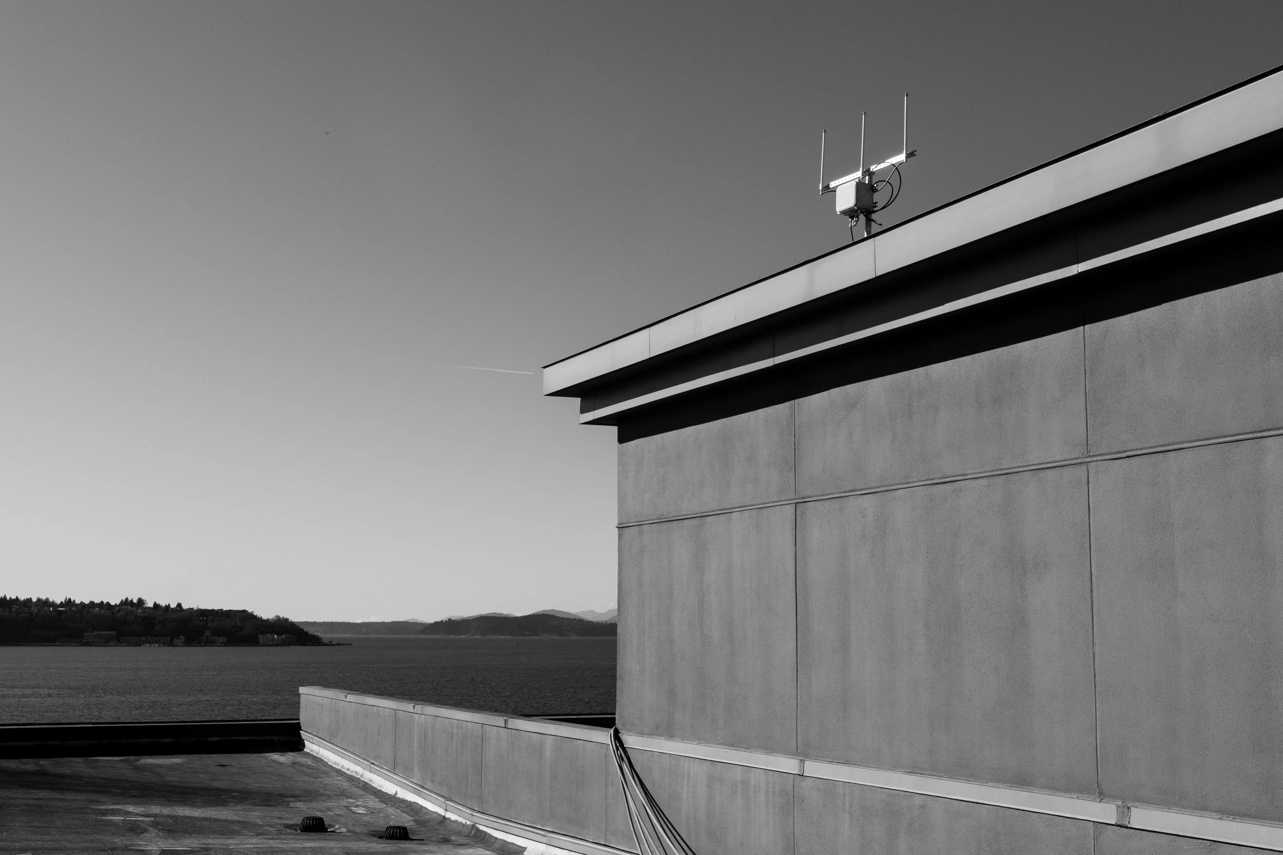 Black and white photo of a modern building with a water view in the background, featuring an antenna on the rooftop.