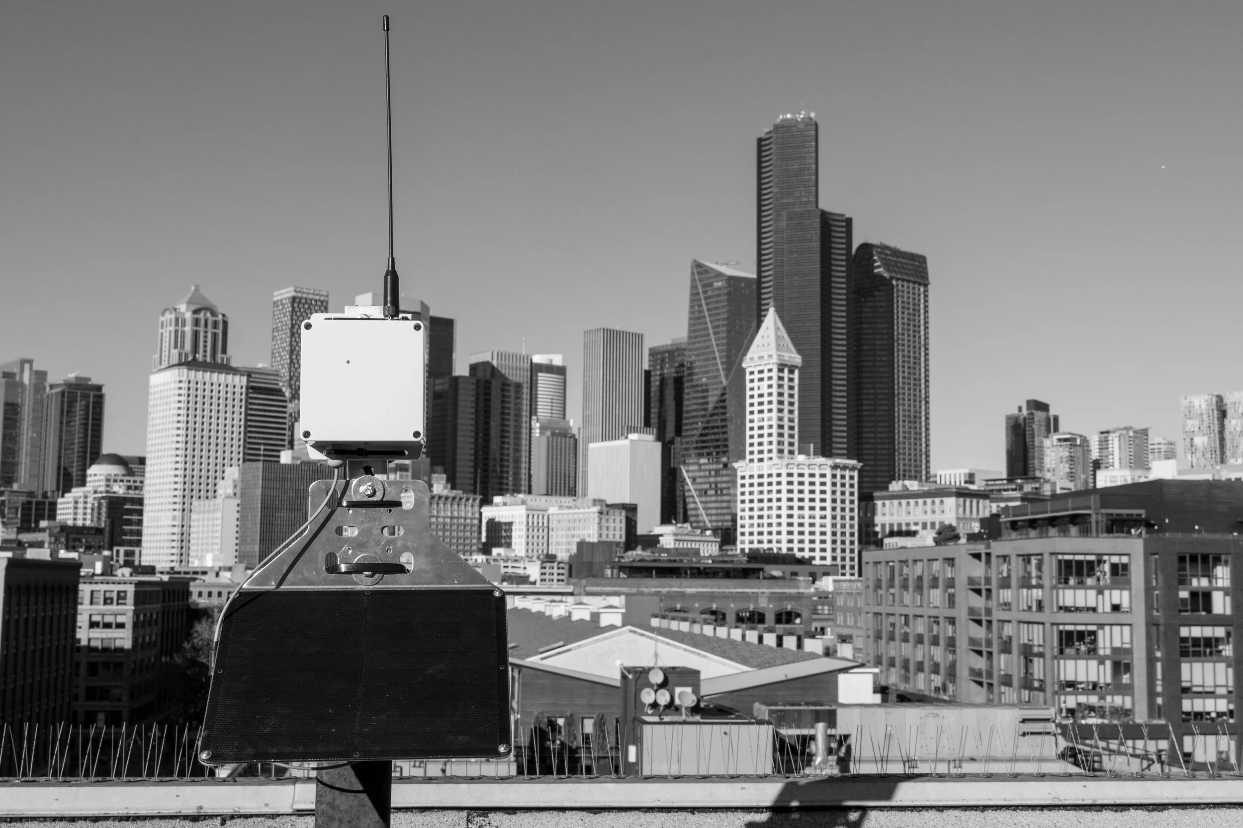 Black and white photo of a city skyline with tall buildings, with an antenna and electronic equipment in the foreground.