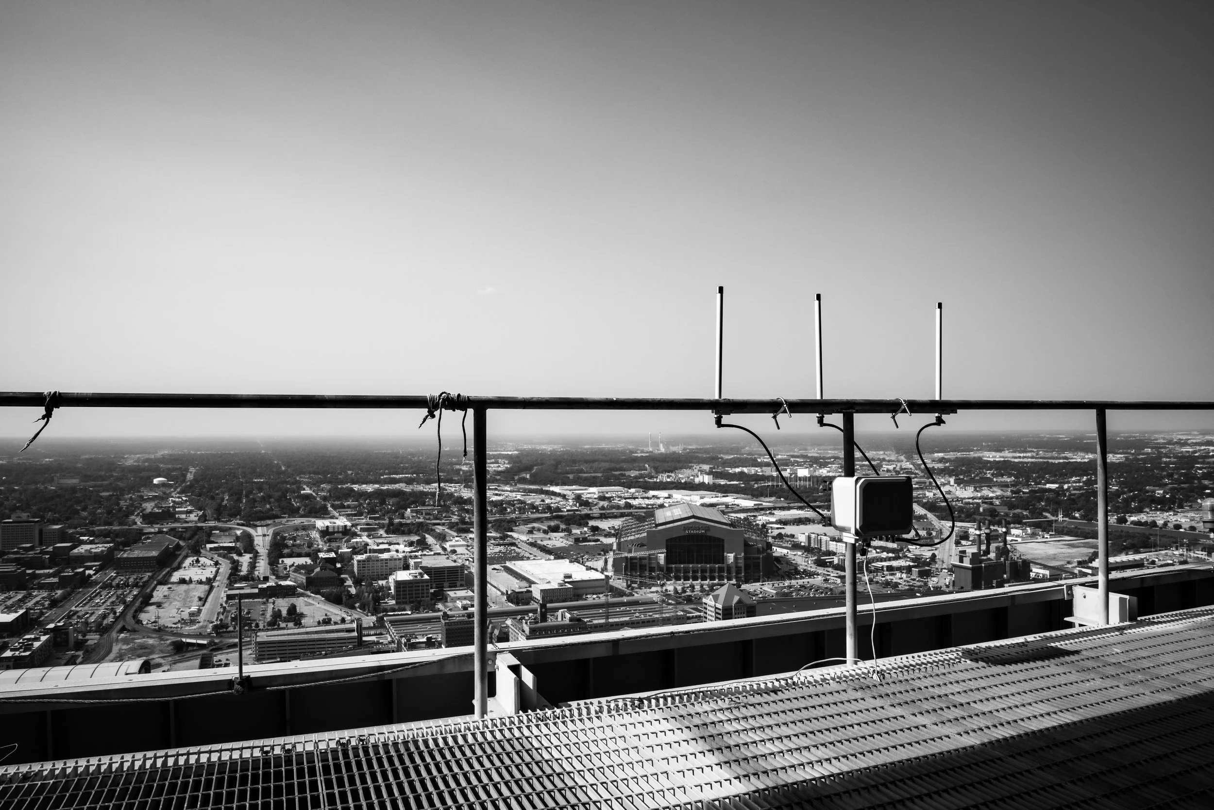 Black and white photo of a city skyline from a rooftop, showing various buildings and roads, with a railing and antennas in the foreground.