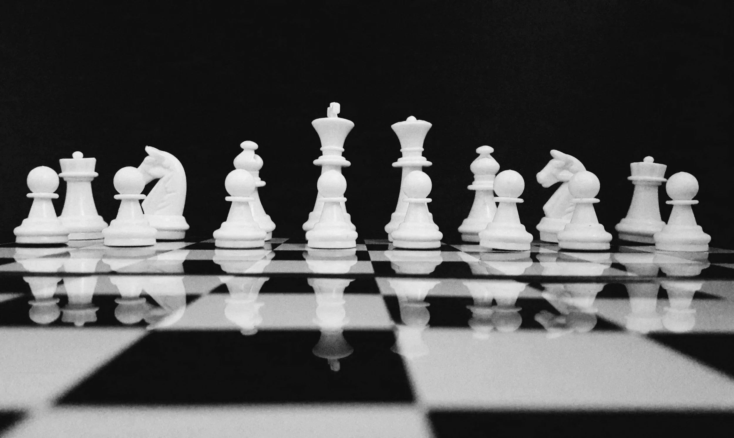 Black and white photograph of a chessboard with white chess pieces arranged in starting positions. The focus is on the pieces, with a mirror-like reflection of the chessboard and pieces visible on the surface below. The background is dark, creating a contrast with the white pieces.