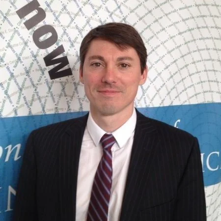 A man in a black suit with a striped tie standing in front of a wall with logo and text.