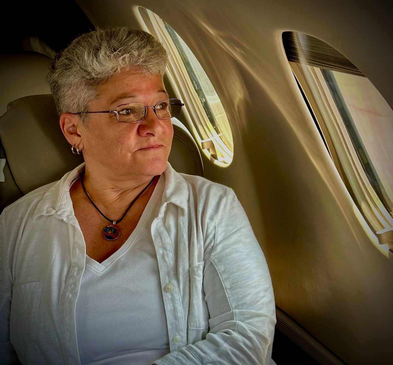 A woman with short gray hair, glasses, and hoop earrings sits by airplane window, looking out.