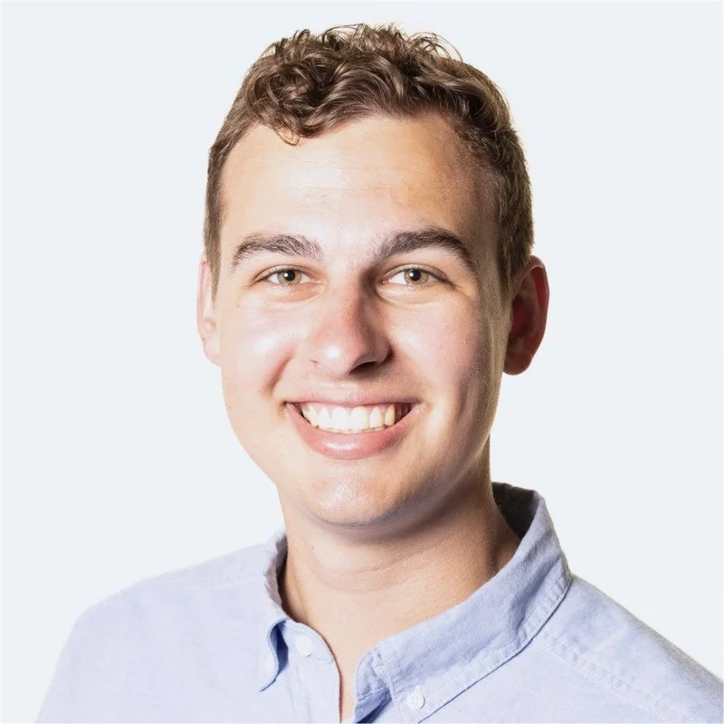Close-up portrait of a young man with curly brown hair, smiling and wearing a light blue collared shirt.