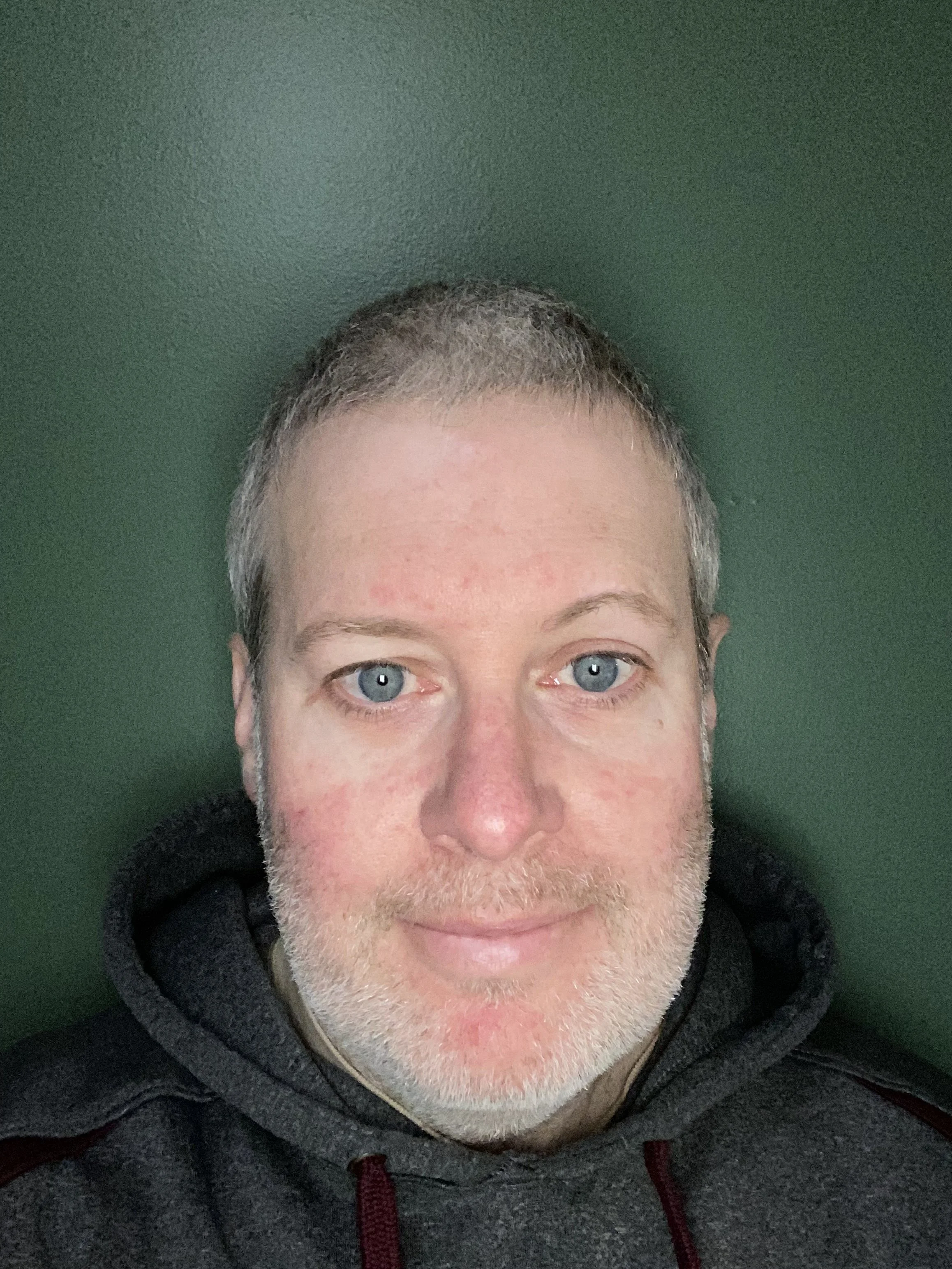 Close-up of a man with light skin, blue eyes, short gray hair, and a beard, wearing a black hoodie, against a green background.