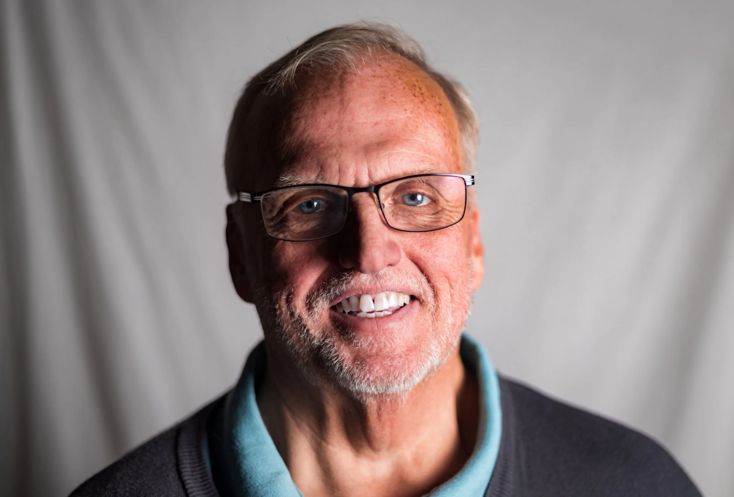 Close-up of a smiling older man with gray hair, beard, and glasses, wearing a black jacket over a light blue collared shirt, against a plain gray background.