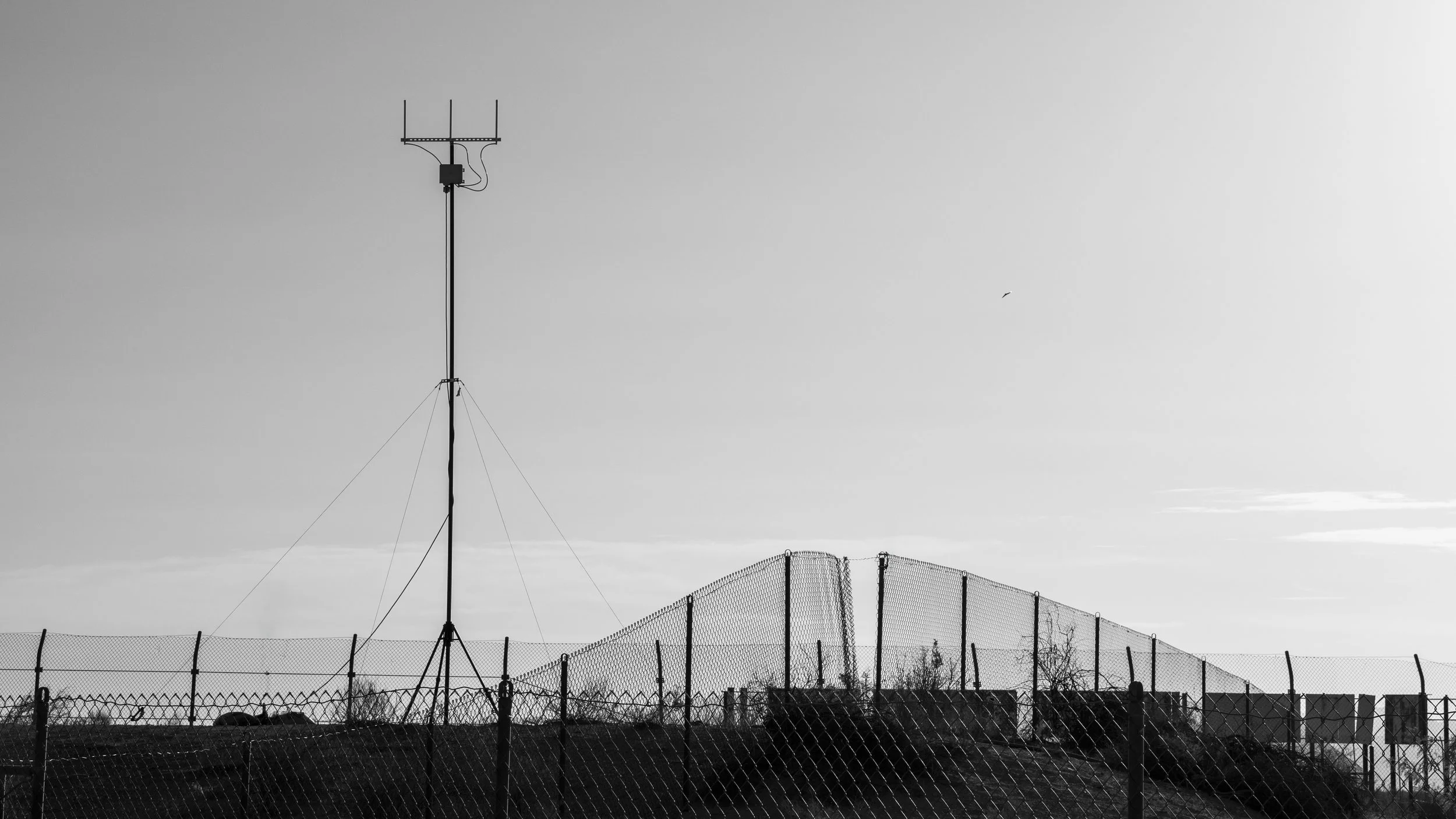 A tall radio antenna tower with a few wires supporting it, set against a partly cloudy sky. There is a chain-link fence in the foreground and some trees and structures in the background.
