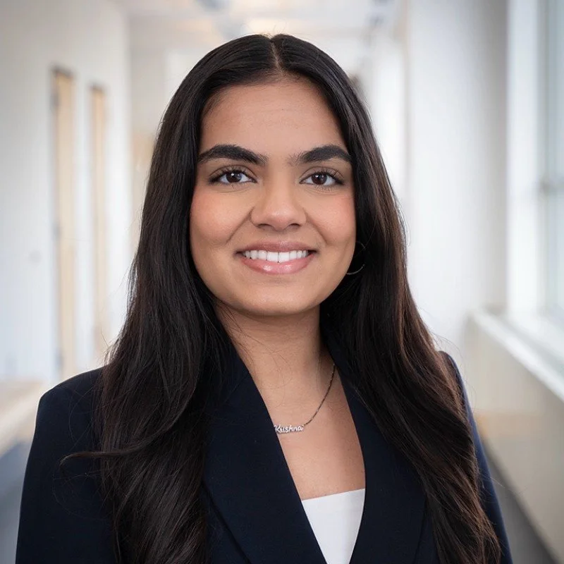 A young woman with long dark hair and a friendly smile, wearing a black blazer and a delicate necklace, standing in a bright office with windows in the background.