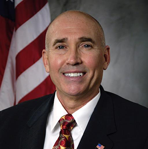 A smiling, bald man in a dark suit and patterned tie standing in front of an American flag.
