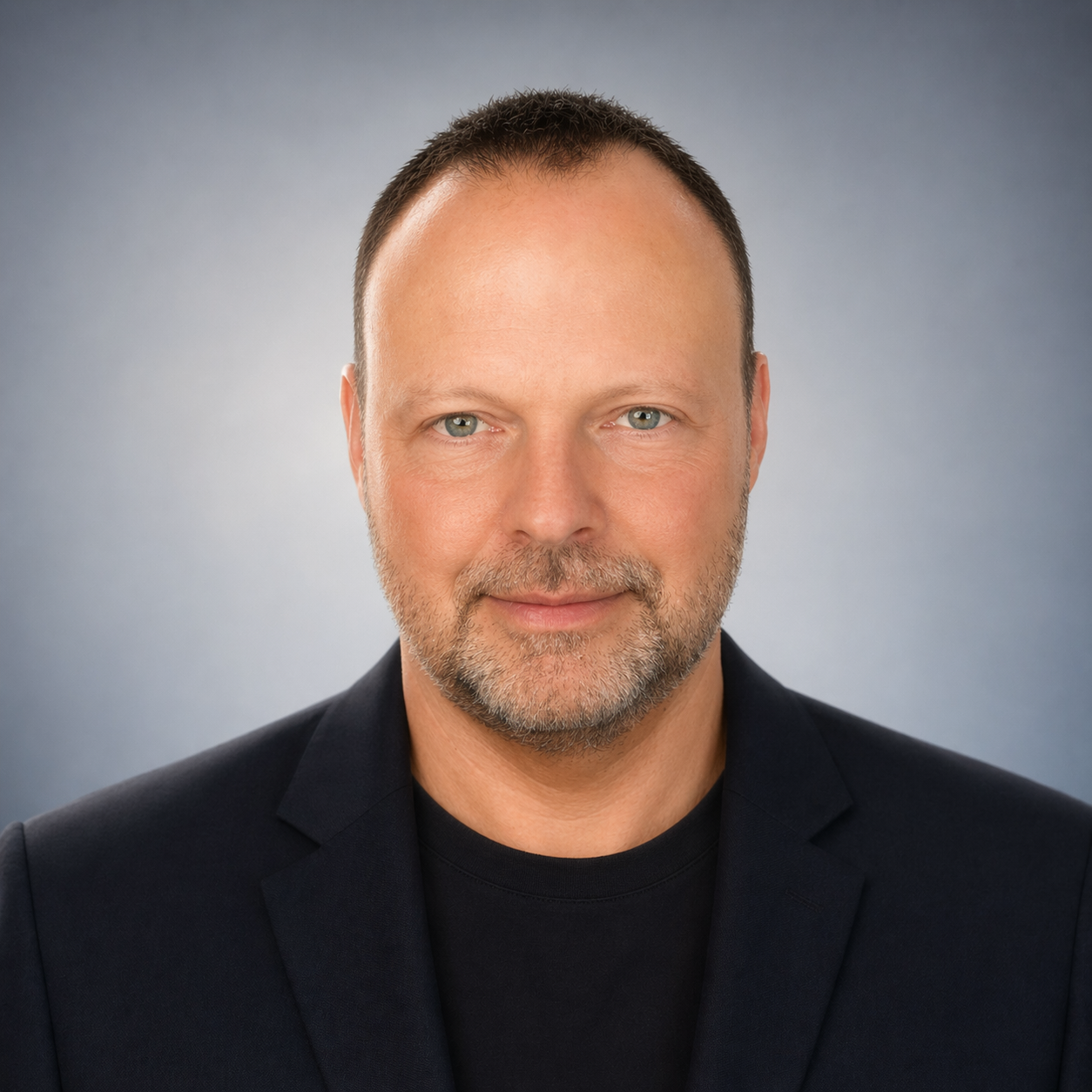 Professional headshot of a middle-aged man with short dark hair, light beard, wearing a dark blazer and black shirt, against a gray gradient background.