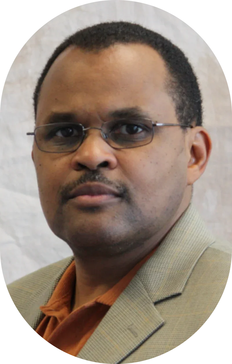 A man with glasses, short black hair, and a mustache wearing a beige blazer over an orange shirt, looking at the camera against a neutral background.