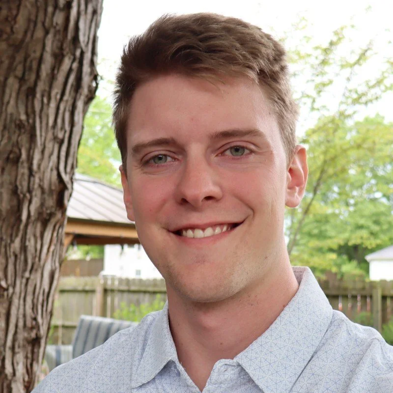 Young man smiling outdoors with a tree and wooden fence in the background.