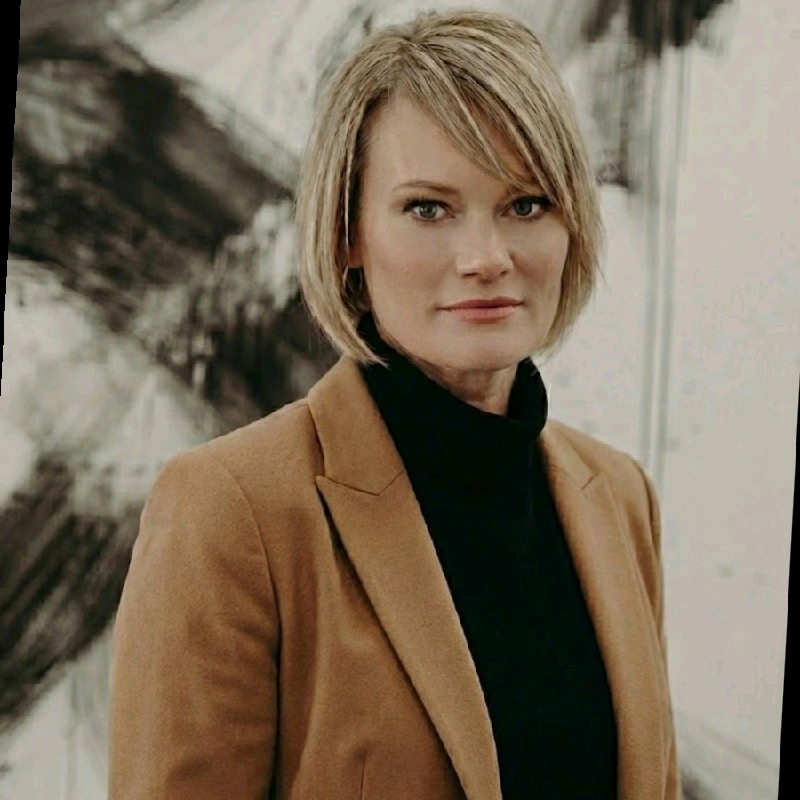 A woman with short blonde hair wearing a camel-colored blazer and black turtleneck, standing indoors in front of an abstract black and white artwork.