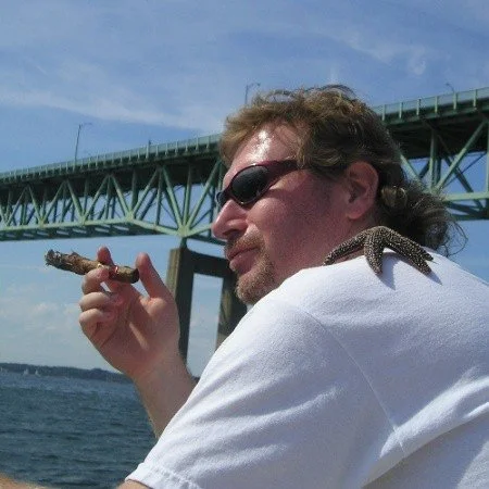 A man wearing sunglasses holding a cigarette with a lizard on his shoulder near a body of water and bridge in the background.
