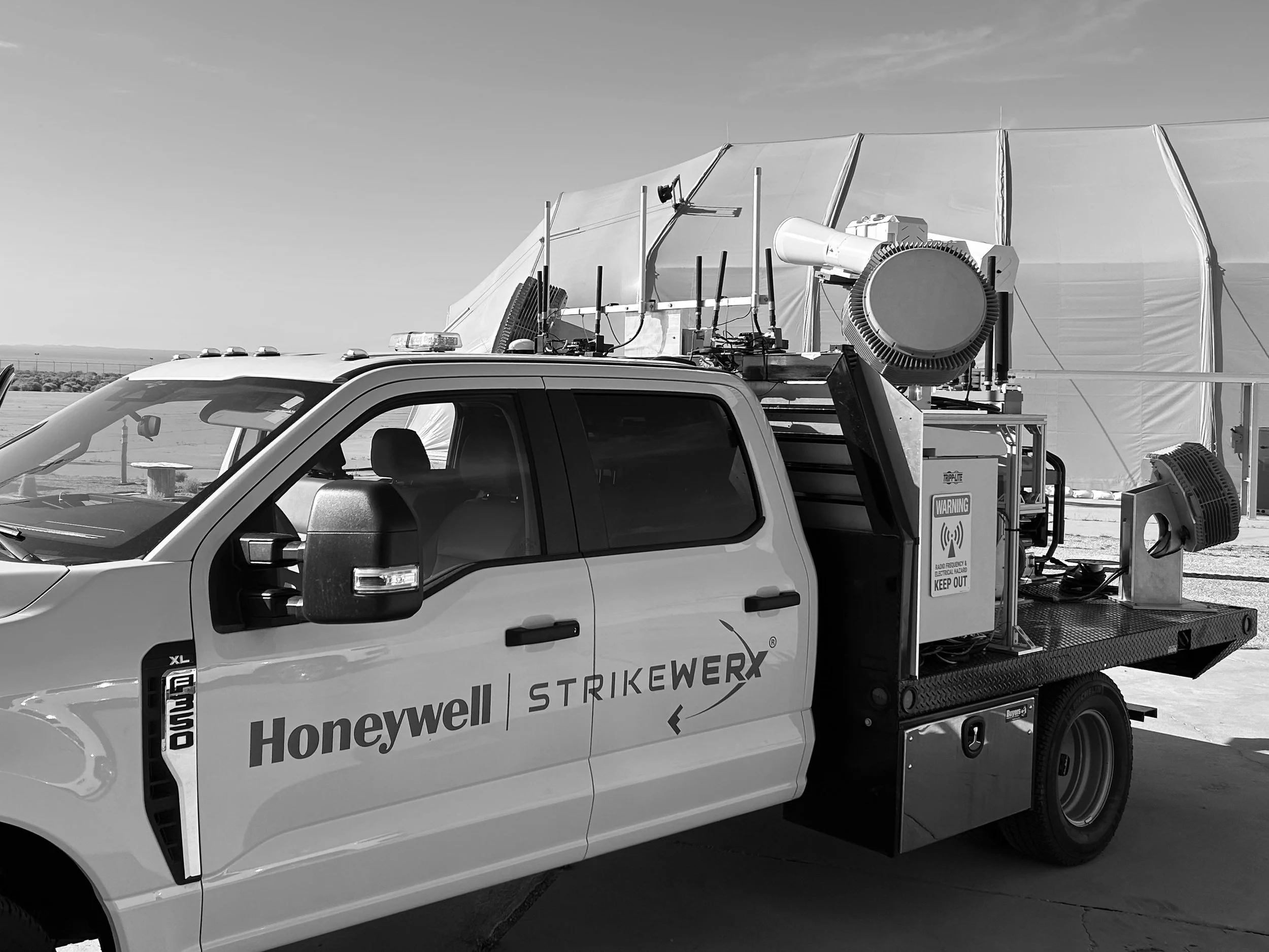 A white utility truck with the words Honeywell and Honeywell Strikewrx printed on the side, equipped with various antennas and equipment on the back, parked outside on a paved area with a large tent and open sky in the background.