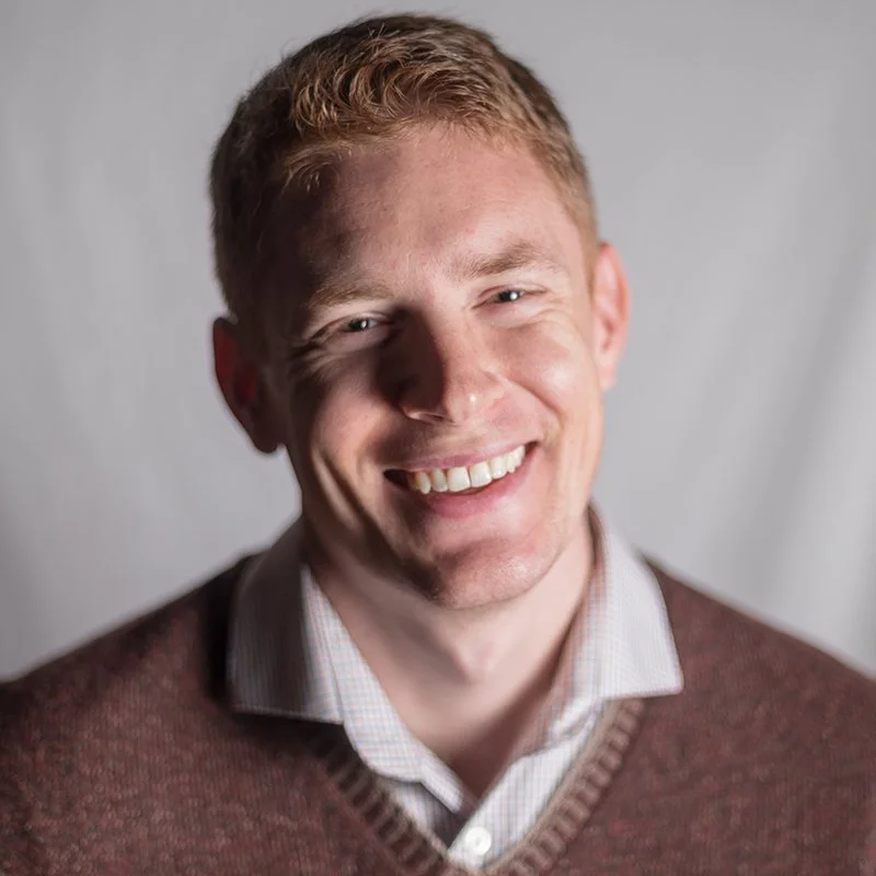 A young man with short red hair and light skin, smiling and wearing a white collared shirt and a brown sweater against a plain gray background.