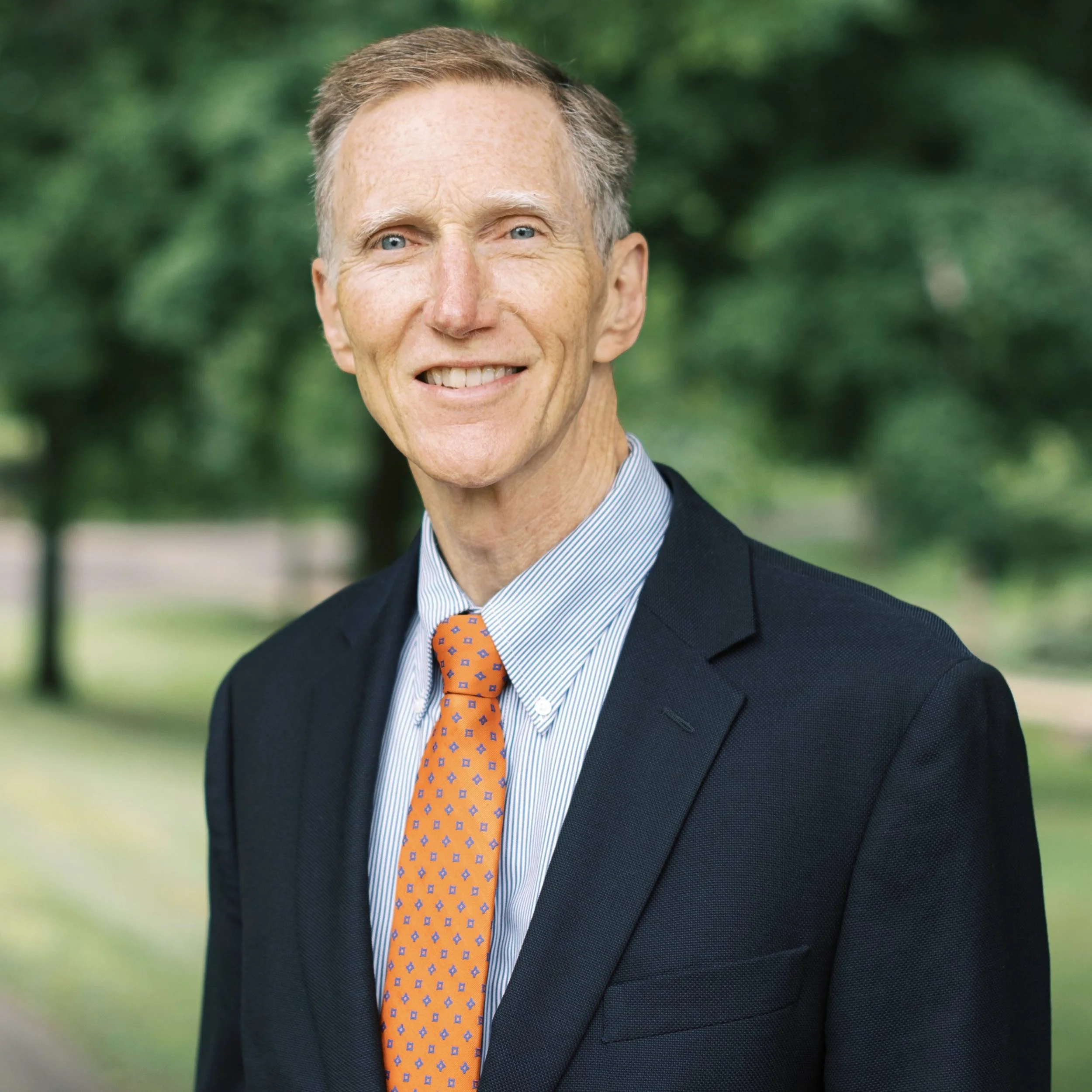 A smiling older man wearing a dark suit, striped shirt, and orange tie, standing outdoors with a blurred green background.