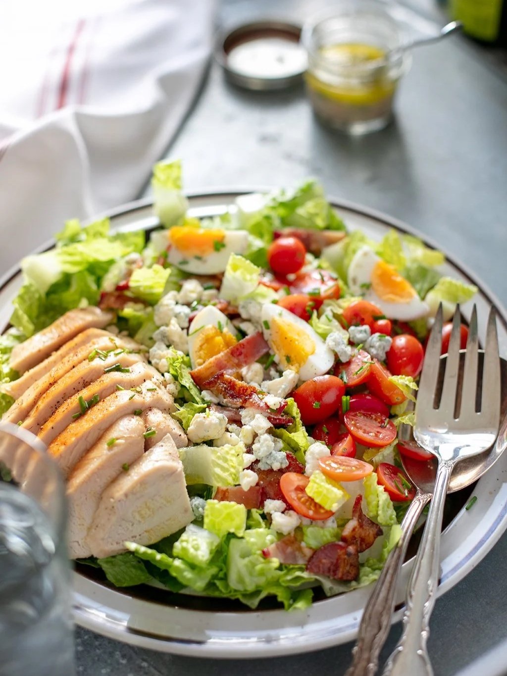 A fresh Cobb salad with chopped lettuce, halved cherry tomatoes, sliced grilled chicken, boiled eggs, blue cheese crumbles, chopped green onions, and pieces of bacon, served on a white plate with silver cutlery.