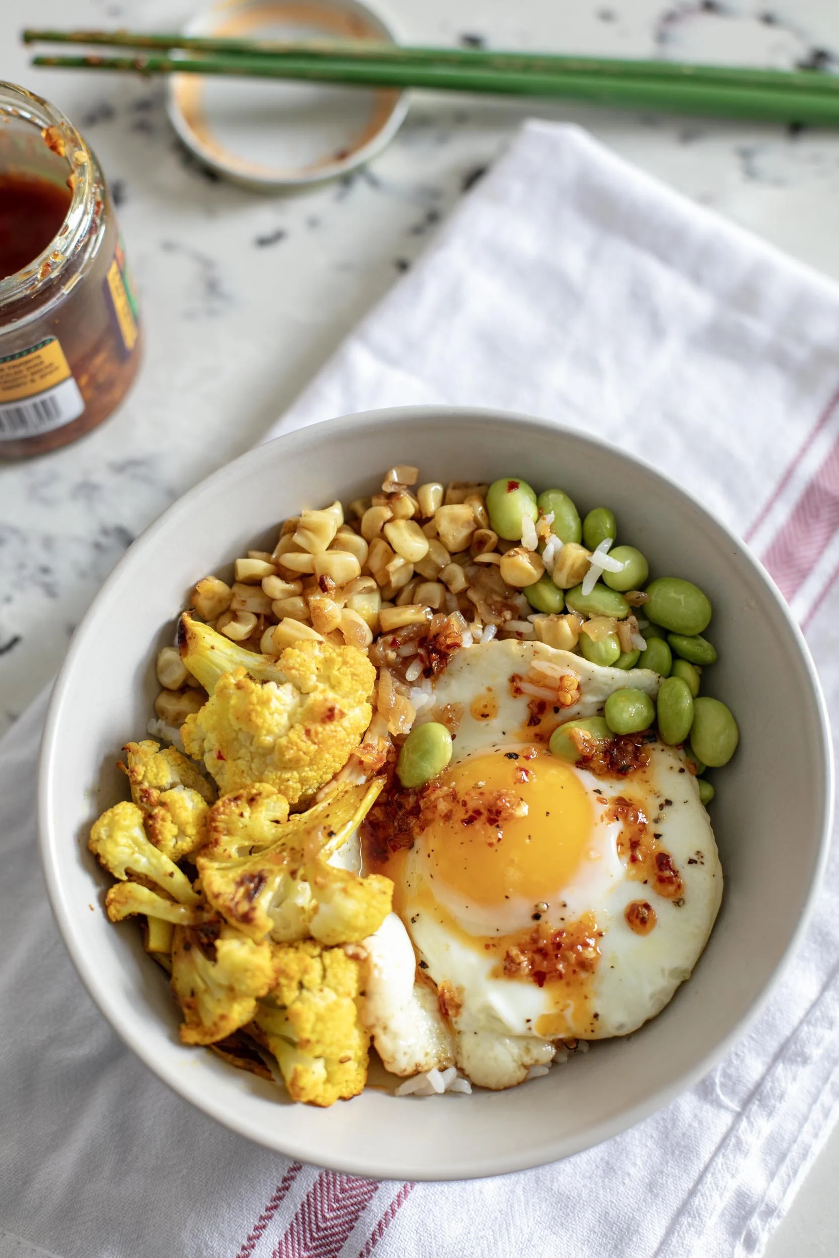 A bowl of breakfast food with cooked cauliflower, edamame, and a fried egg.