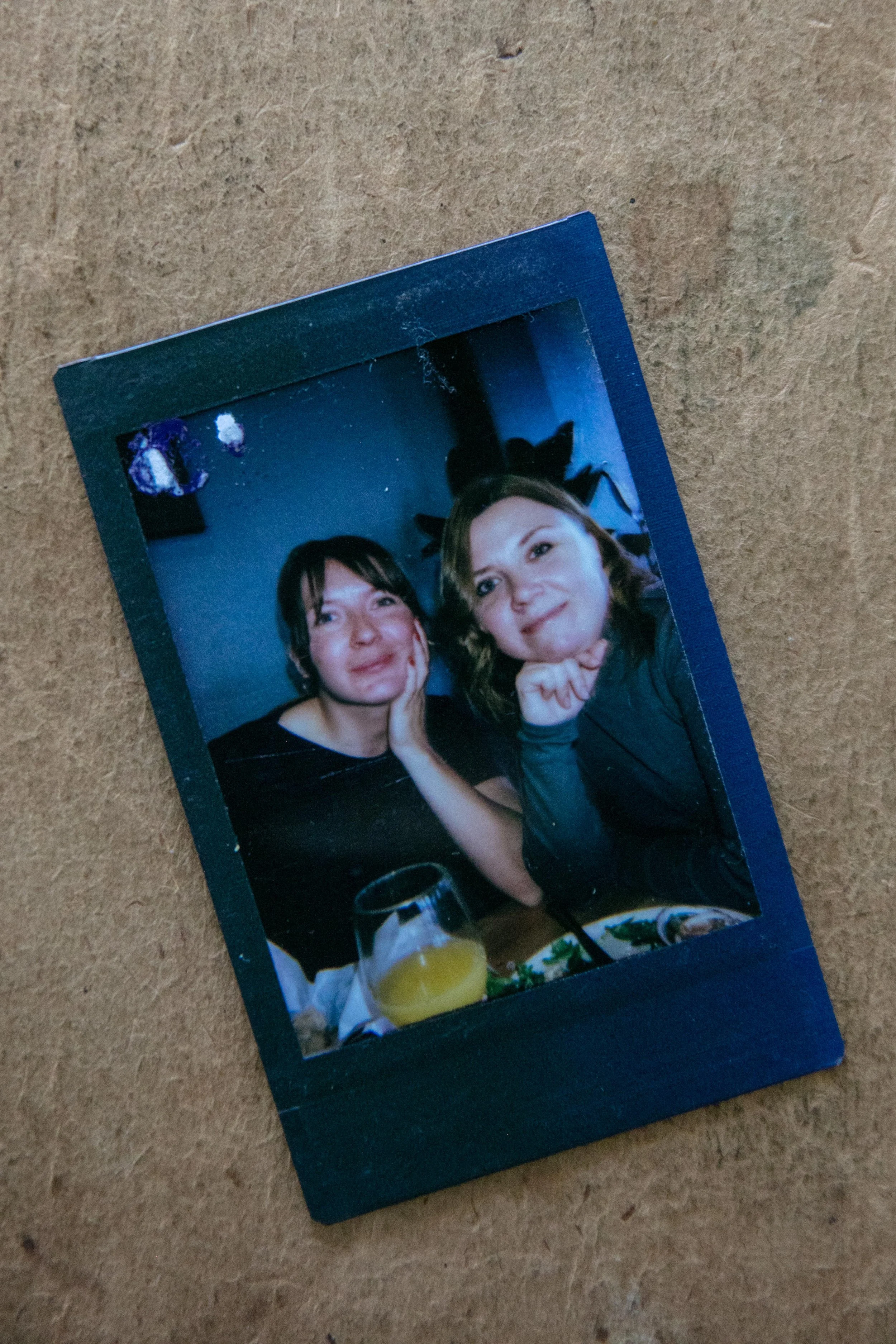 A photograph of a printed picture photo featuring two women sitting together at a table, with various drinks and items visible on the table, and some plants and decorations in the background.