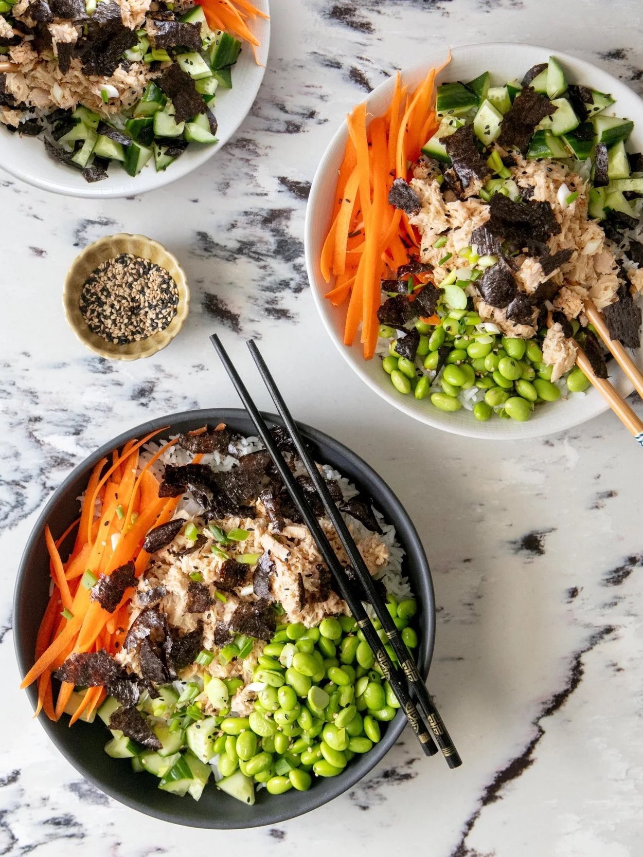 Bowl of Asian-inspired rice bowls with shredded chicken, shredded carrots, chopped cucumbers, edamame, seaweed strips, and chopped green onions, with chopsticks on top.