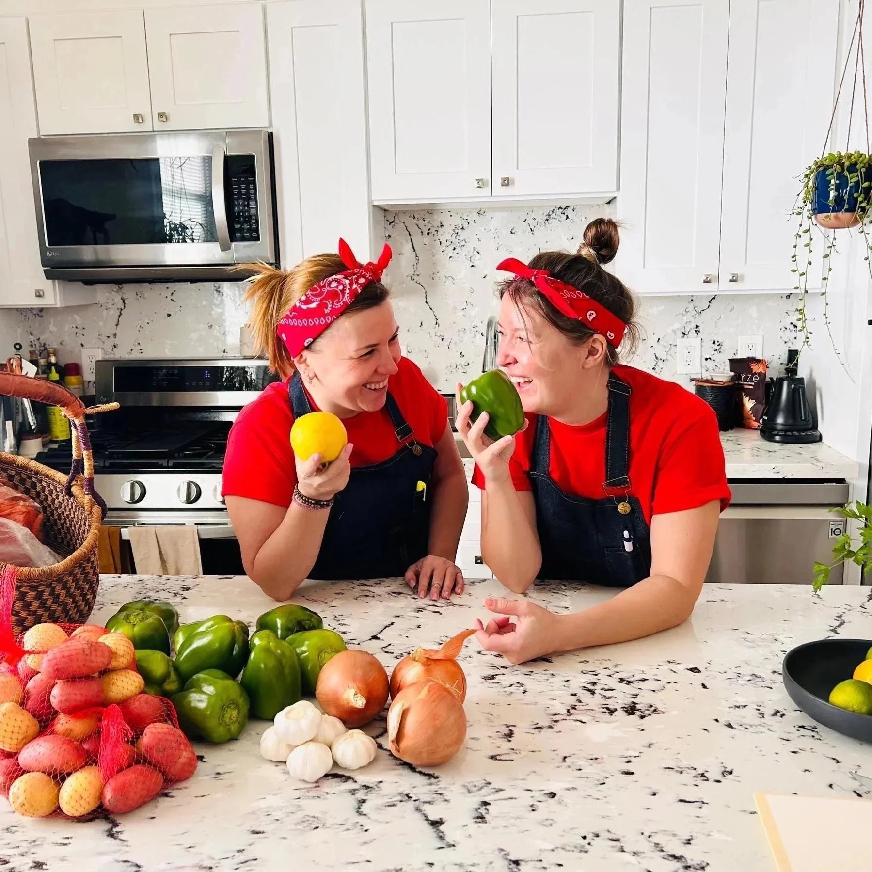Two women wearing red shirts and red bandanas on their heads are smiling and holding colorful bell peppers in a kitchen. The kitchen has white cabinets, a marble countertop, and various vegetables including onions, garlic, and potatoes on the counter.