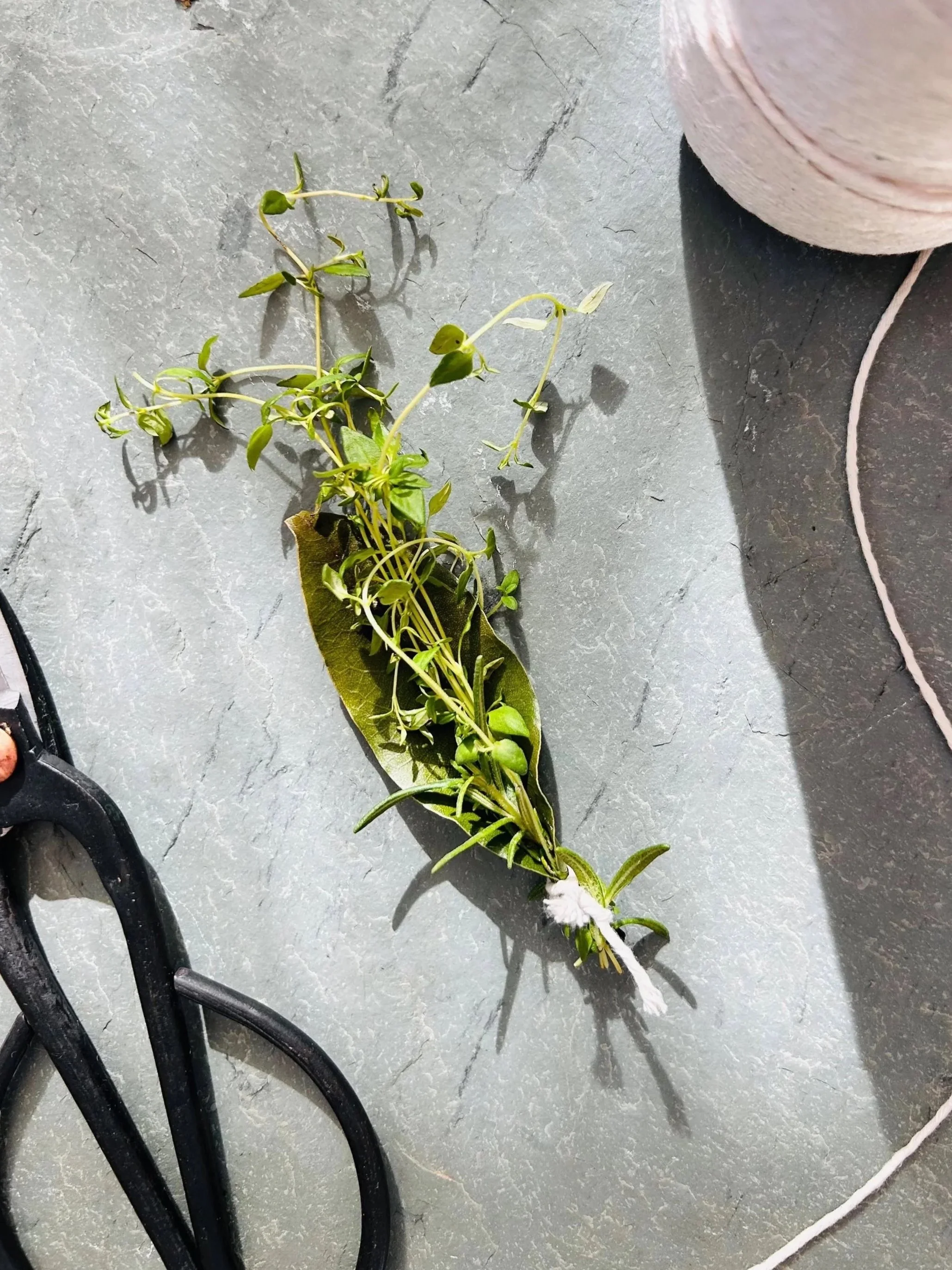 A small bunch of fresh herbs, possibly thyme, tied together with white string, lying on a gray textured surface. Part of a pinkish-gray yarn or thread spool and a black object are also visible nearby.