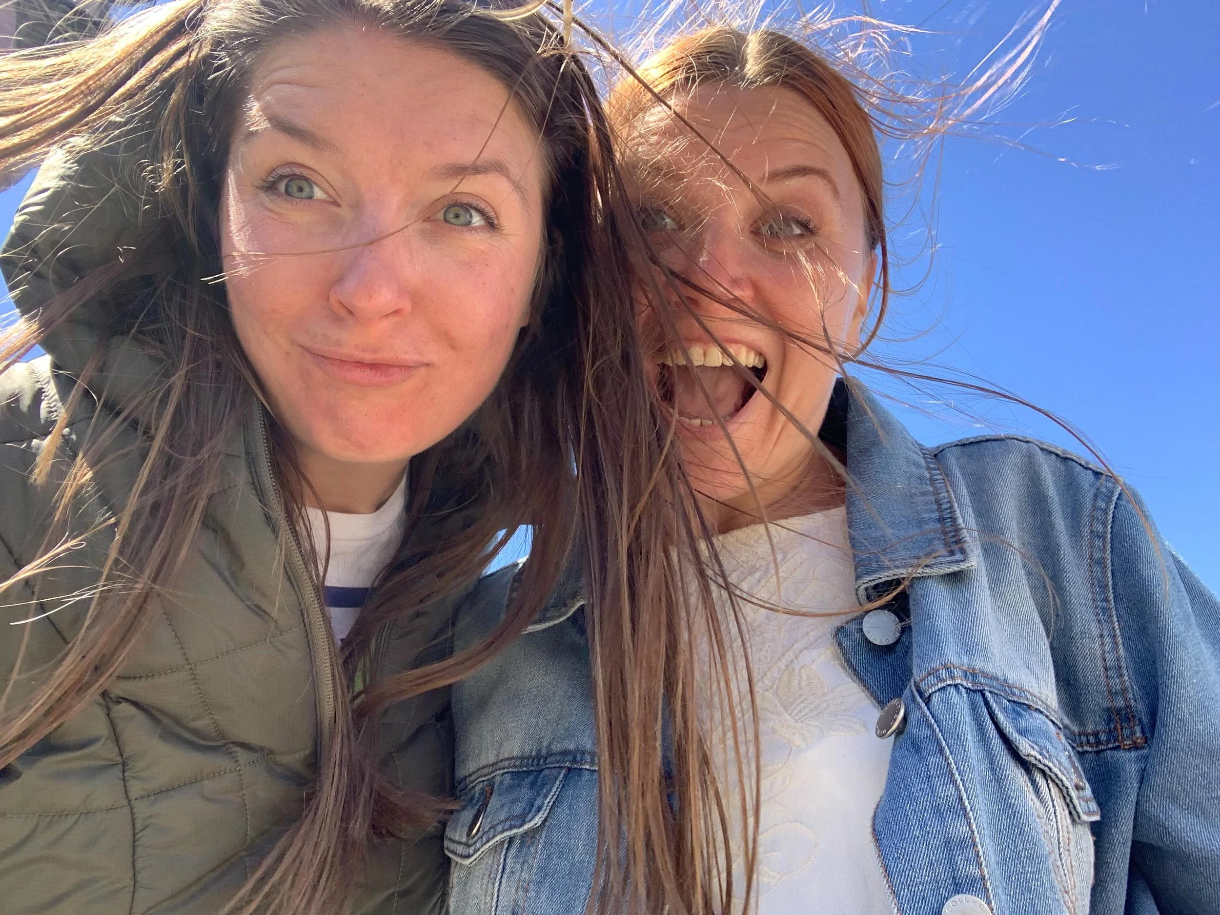 Two women with windblown hair take a selfie outdoors against a blue sky, smiling and looking at the camera.