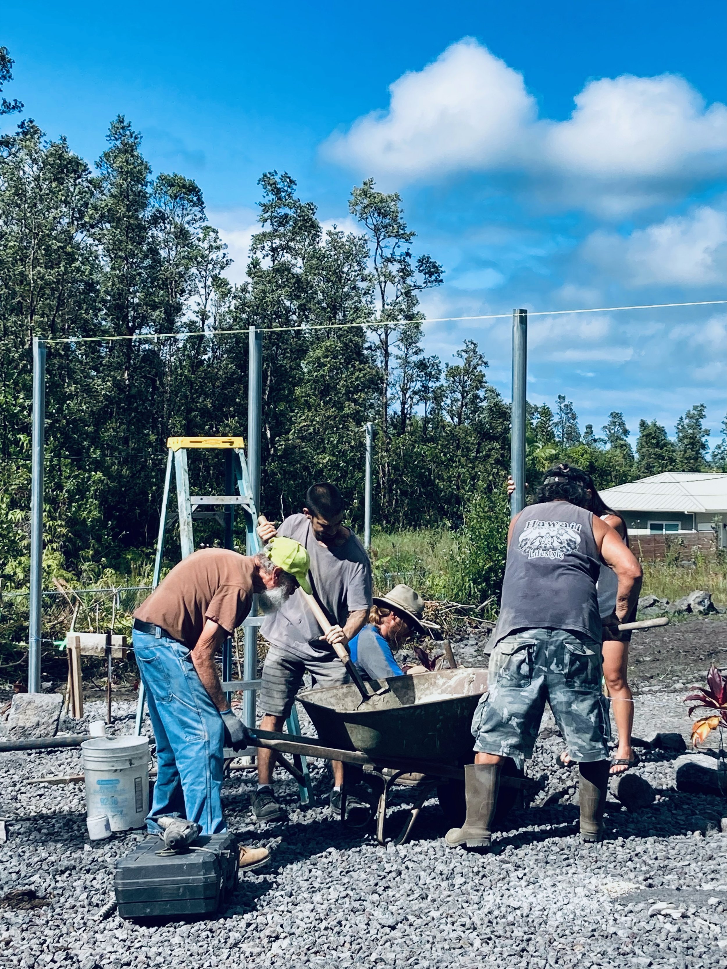 People working together on a construction project outdoors, with trees, a blue sky, and some clouds in the background. They are handling tools and equipment.
