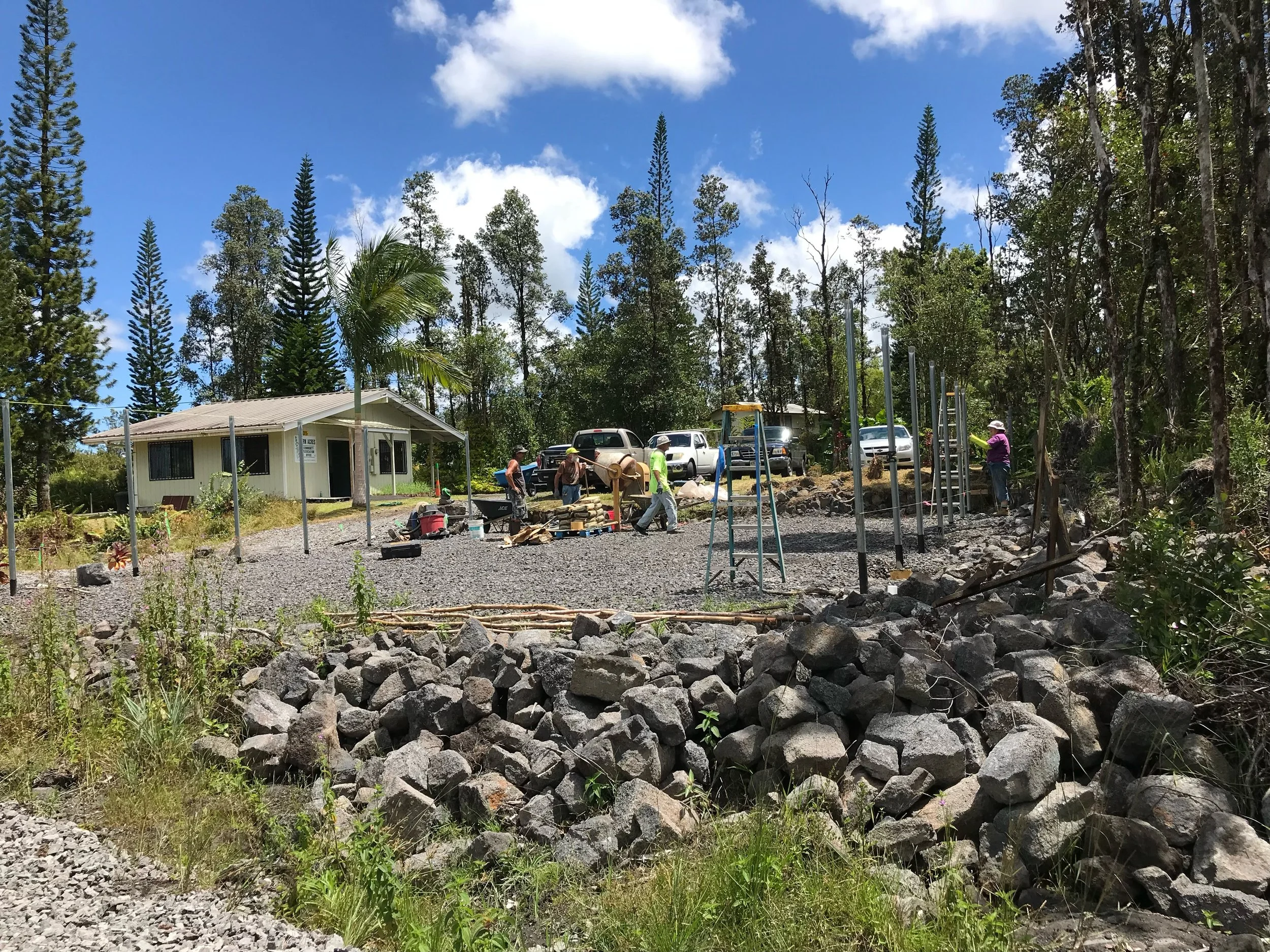 Construction workers working on a building site with a small beige building, trees, and a blue sky with clouds in the background.