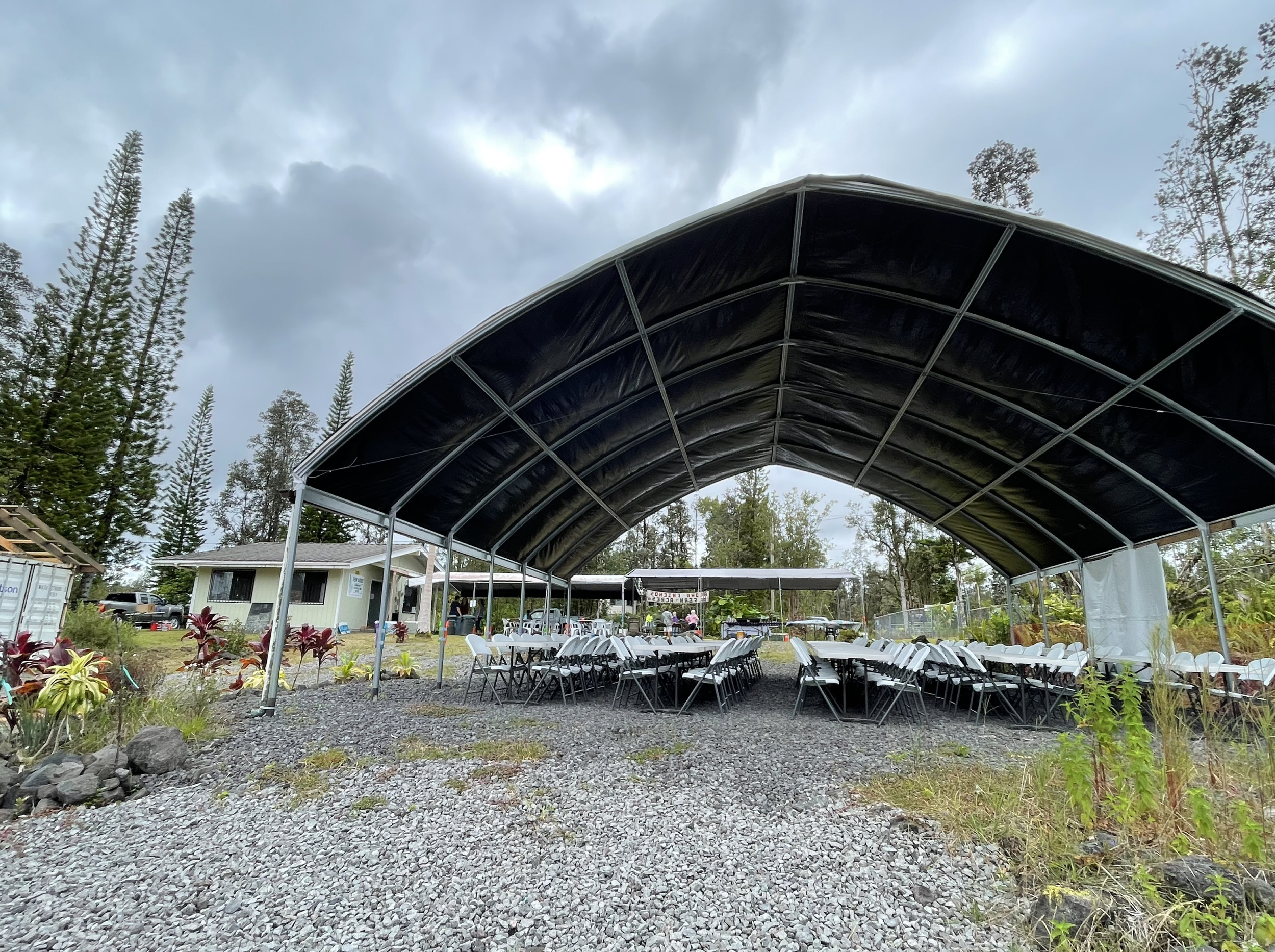 An outdoor event setup with rows of white chairs under a large black canopy, surrounded by trees and a cloudy sky.