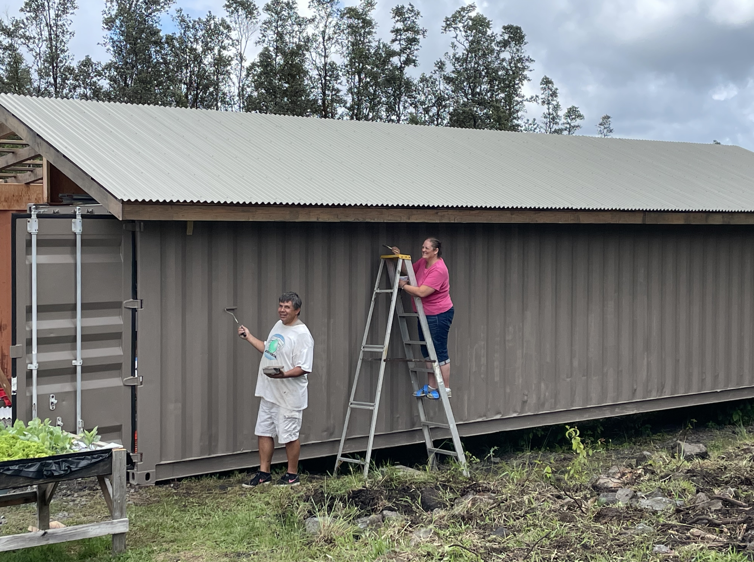Two people installing a large corrugated metal roof on a small building. One person is standing on a ladder, holding tools and working on the roof. The other person is standing on the ground, smiling and holding a tool. The building is gray with vert