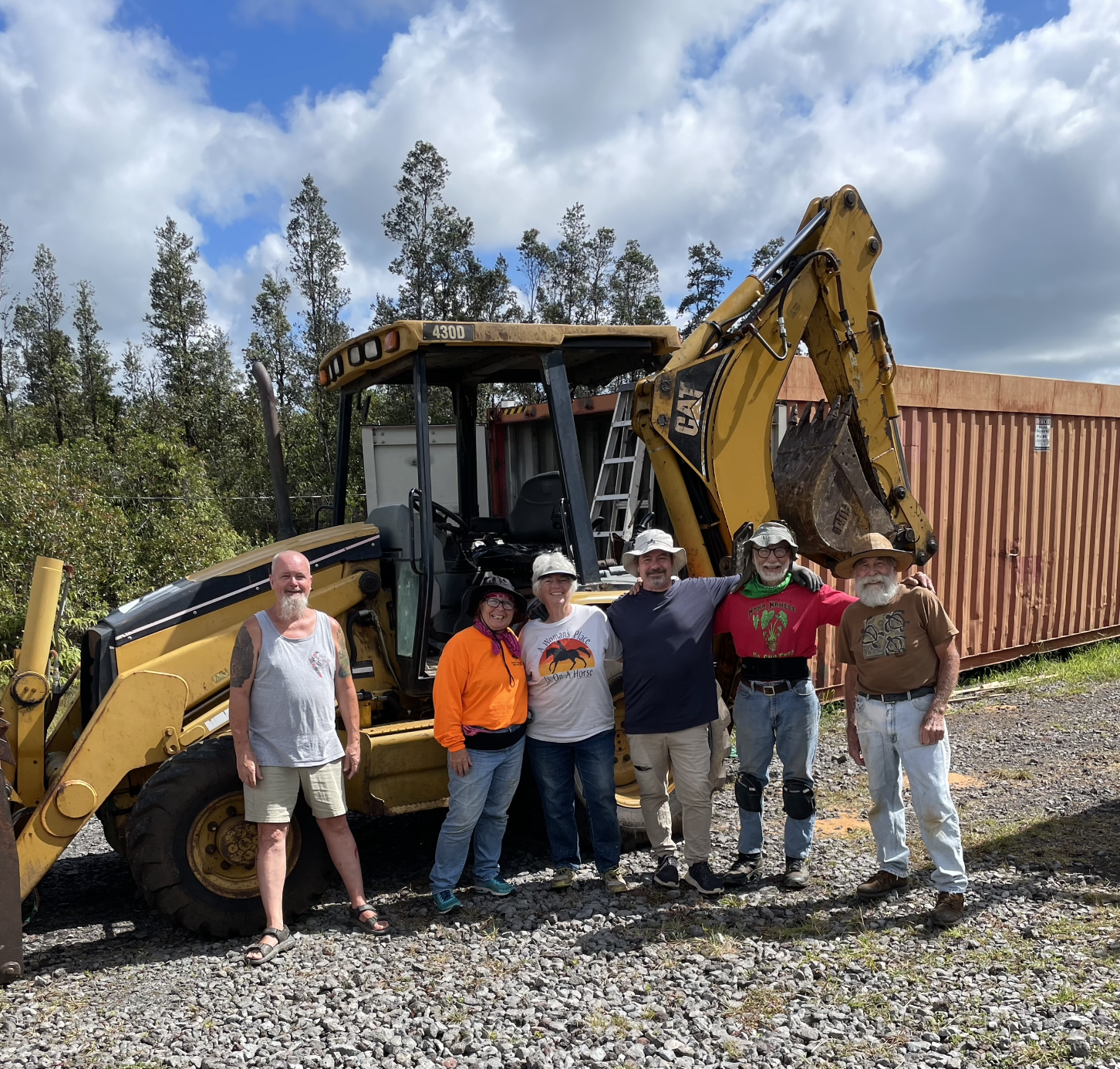 Six people standing in front of a large yellow backhoe loader, smiling at the camera. They are dressed casually, with some wearing hats. Behind them is a large orange shipping container and trees under a partly cloudy sky.