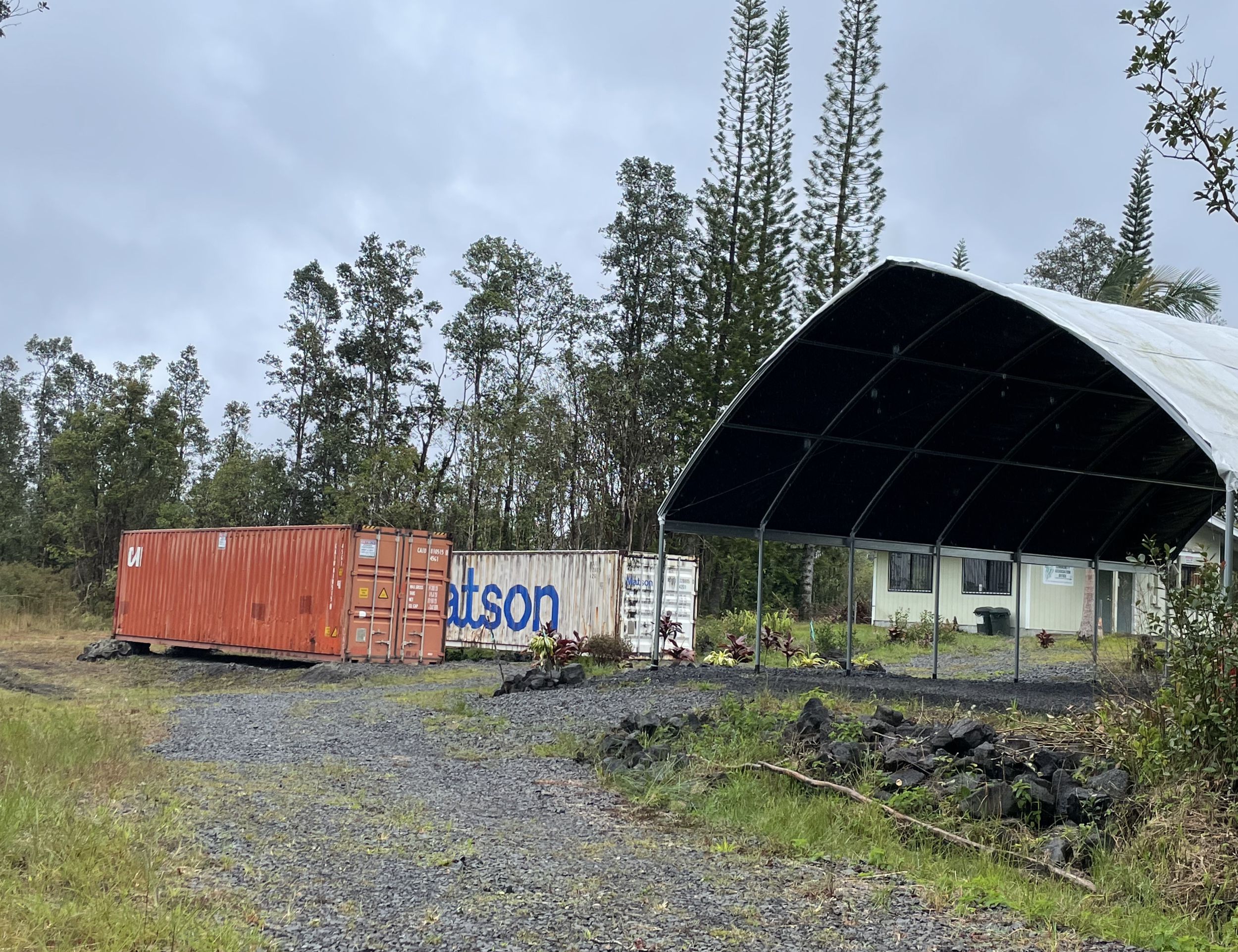 A rural scene featuring a large black covered structure with an arched roof, trees in the background, and shipping containers on the ground in front of the structure, with cloudy sky overhead.