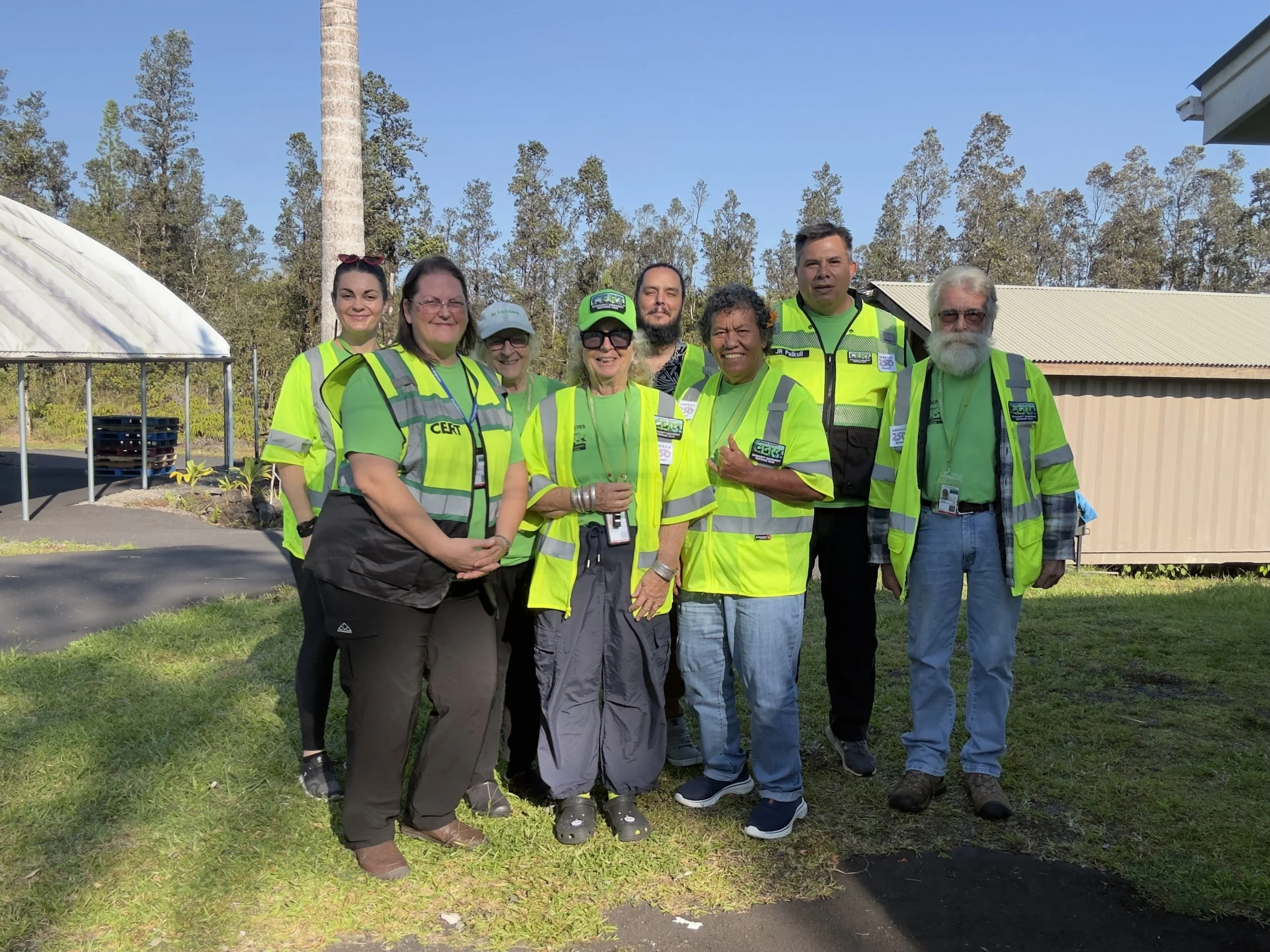 Group of eight diverse people wearing bright yellow safety vests outdoors, standing on grass with trees and a building in the background, smiling at the camera.