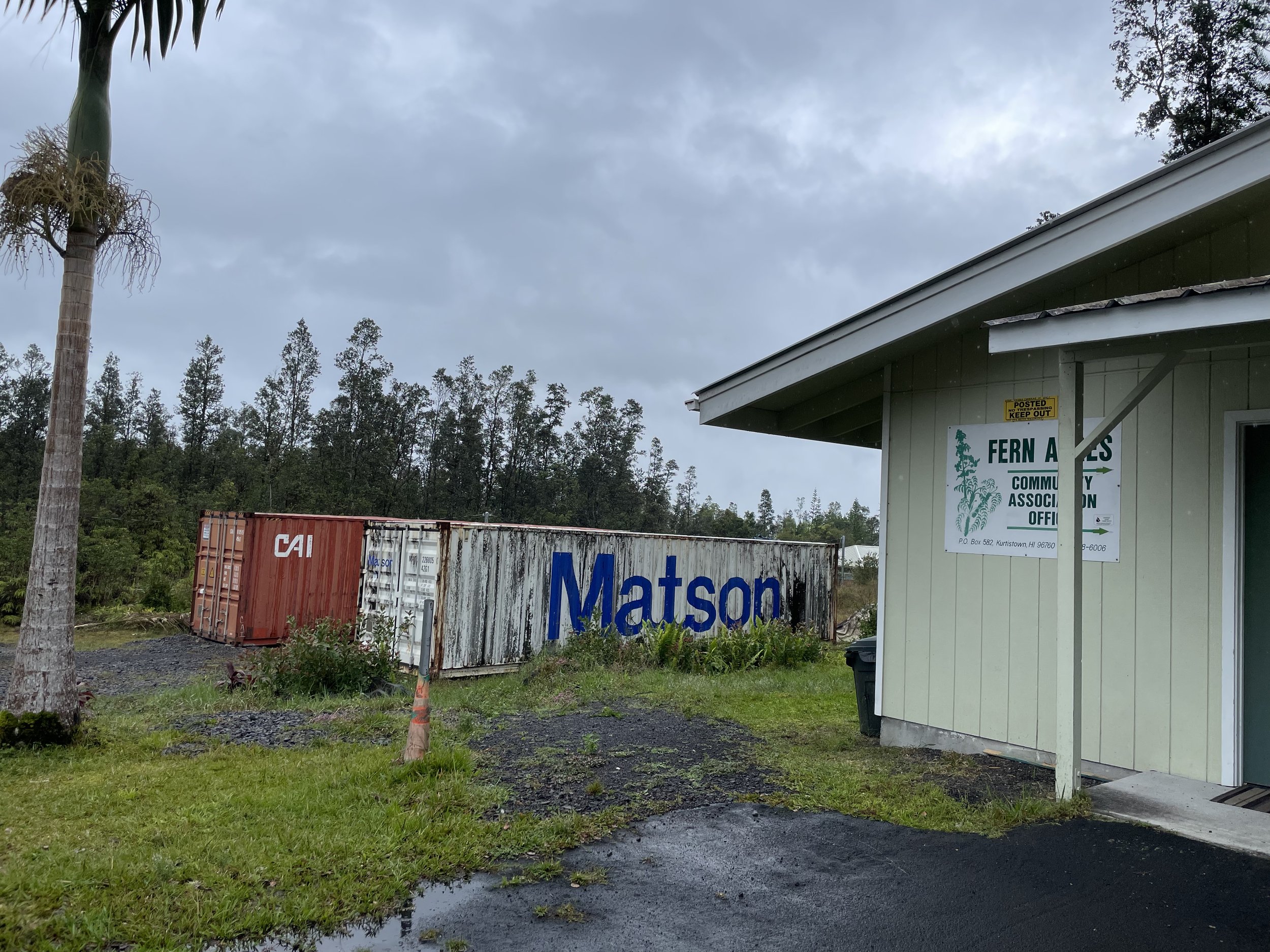 Overcast sky with gray clouds, trees in the background, a small building with a sign for Fern Acres Community Association, and colorful shipping containers in the foreground, including a red one labeled 'CAI' and others with branding.