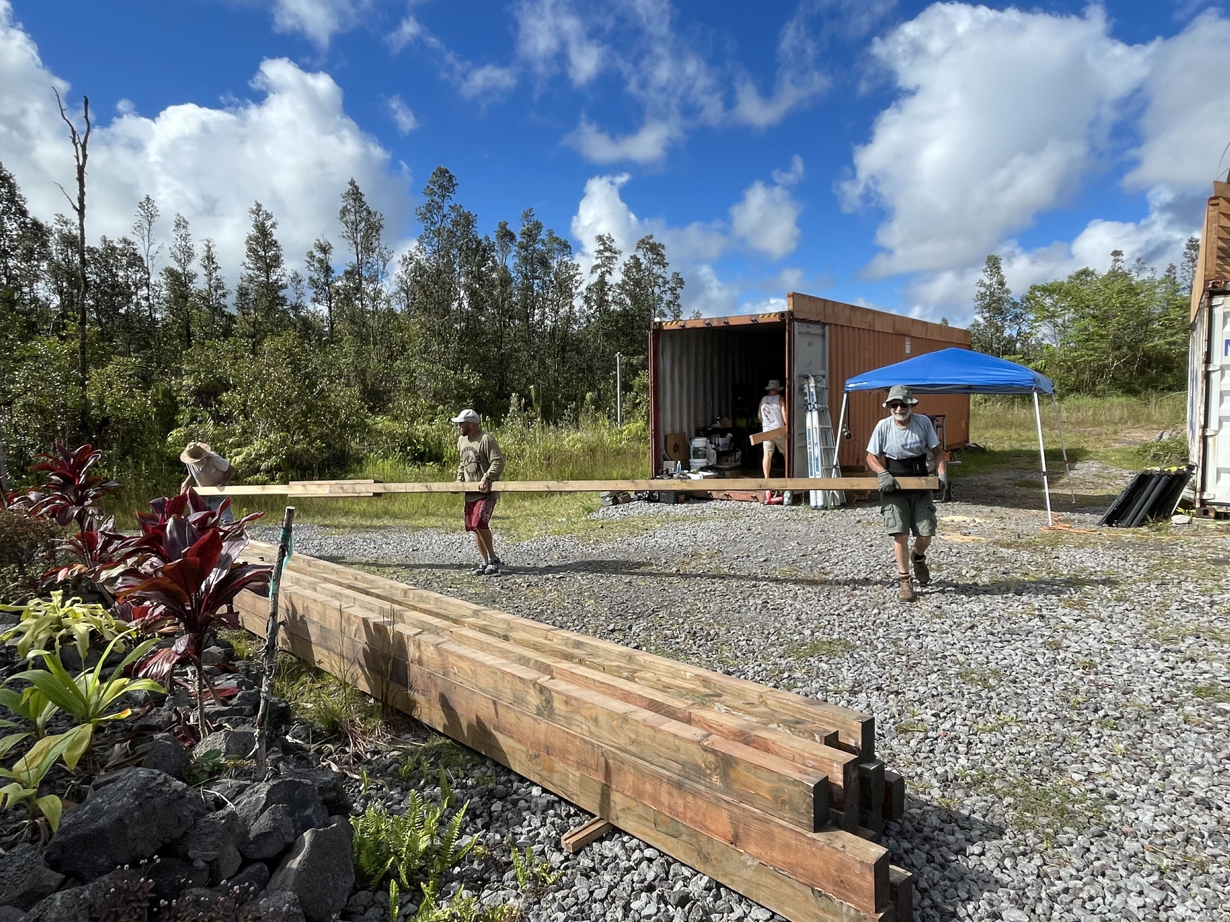 People working outdoors, building a wooden structure in a grassy area with trees and a blue sky with clouds.