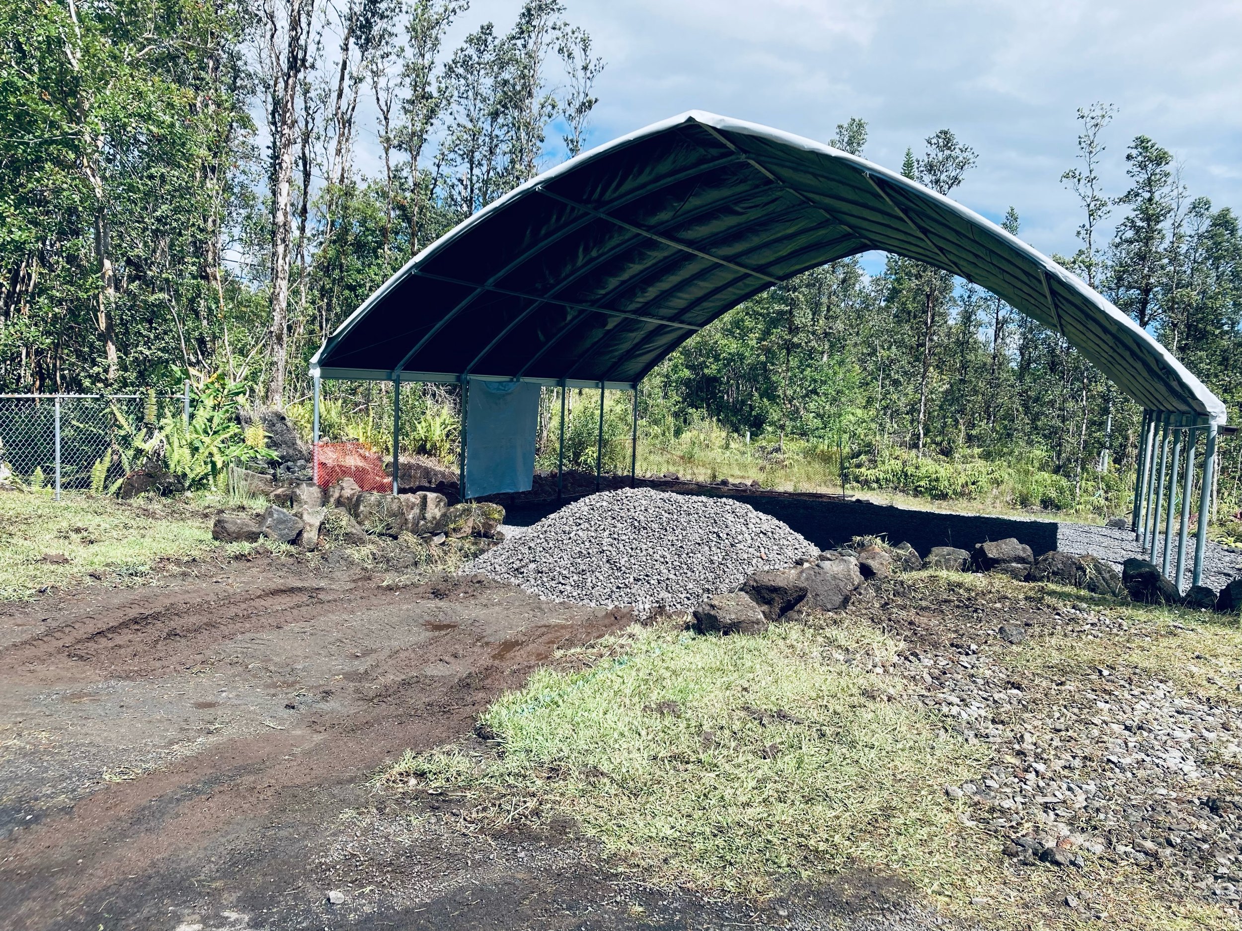 A large, partially constructed metal shelter with a curved roof in a rural, wooded area, surrounded by trees, a chain-link fence, and uneven ground with rocks and dirt.