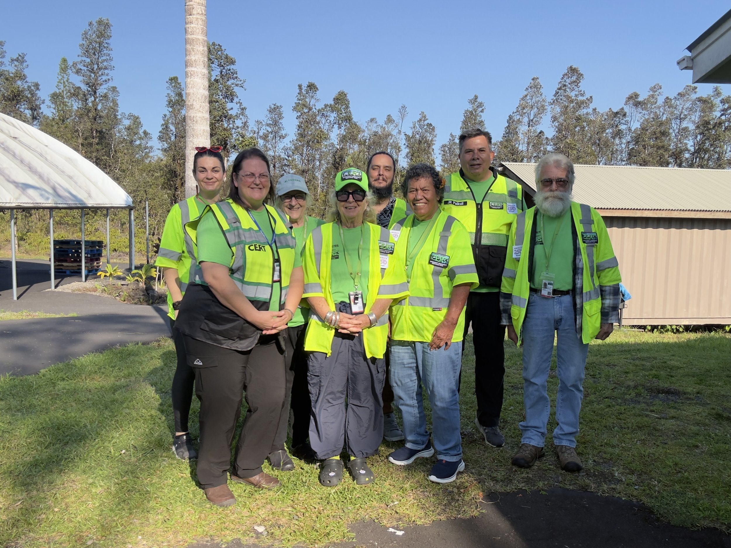 A group of nine people wearing bright yellow safety vests standing outdoors on a grassy area with trees and a building in the background.