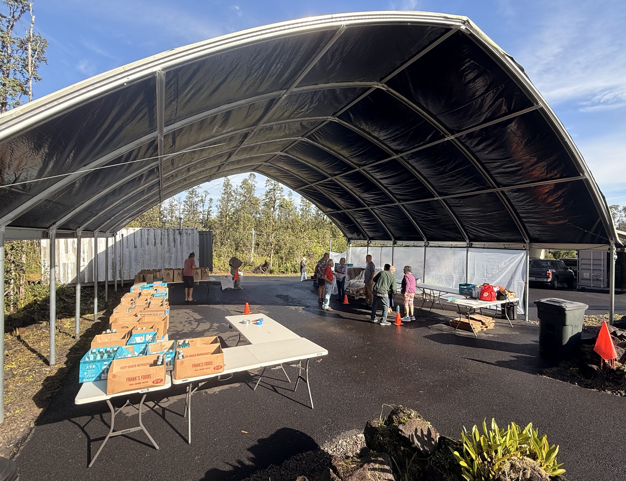 People gathering under a large, open, black-tarpaulin canopy at a community event with tables and boxes of supplies.
