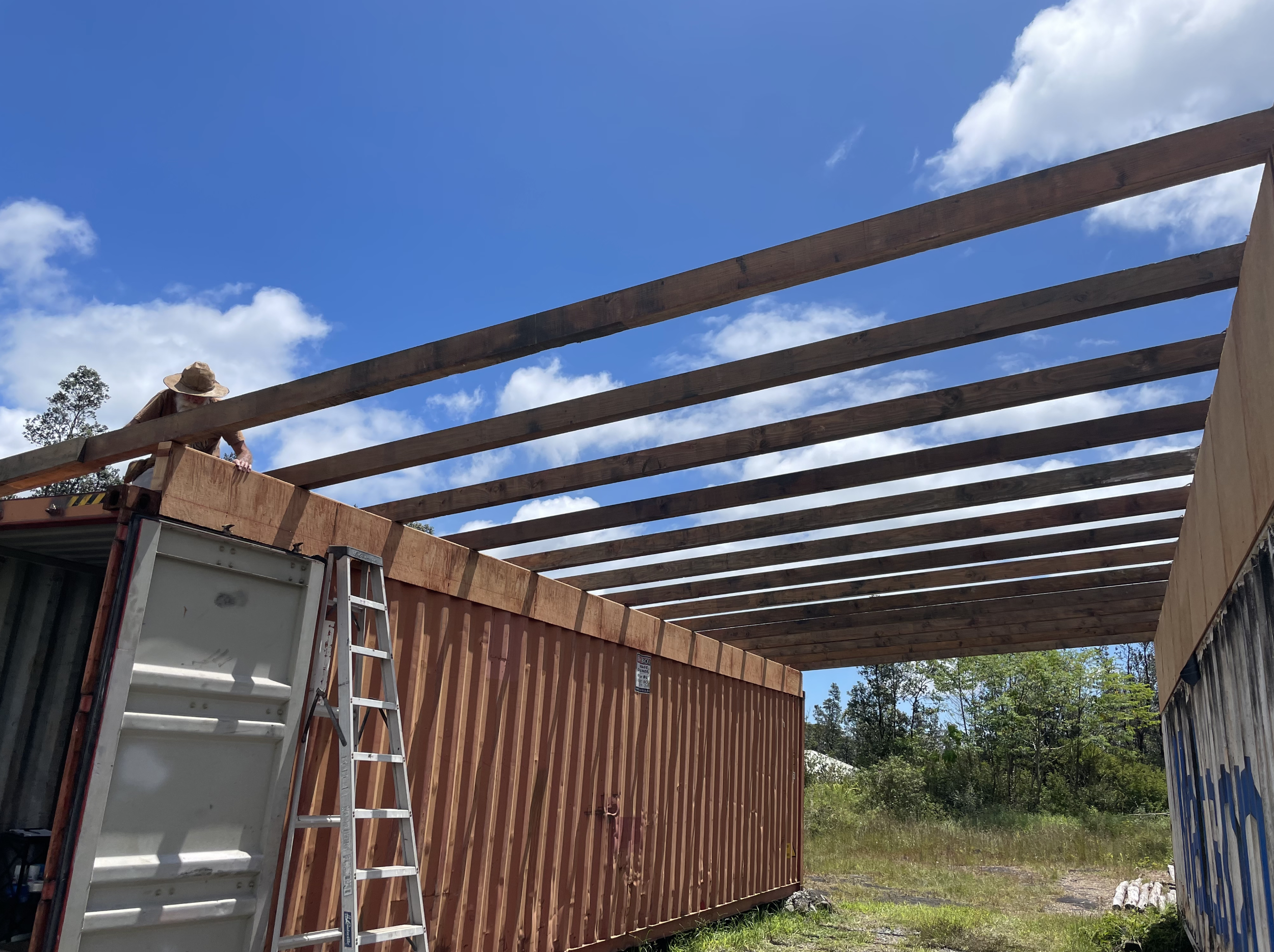 Construction site with wooden beams for building a roof, ladder, and a worker wearing a hat on a truck. Clear blue sky with some clouds and trees in the background.