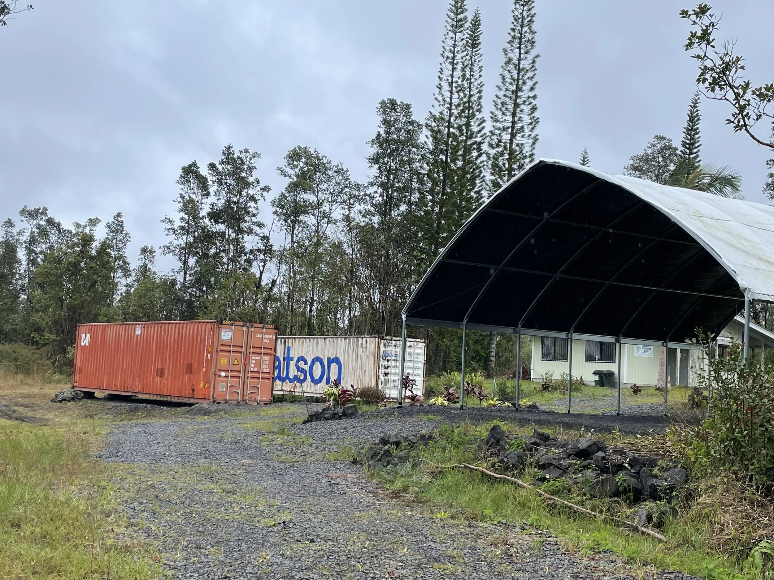 A large orange shipping container with white and blue lettering is situated on a gravel driveway in front of a house with a metal carport. Behind the container are tall pine trees and dense greenery under a cloudy sky.