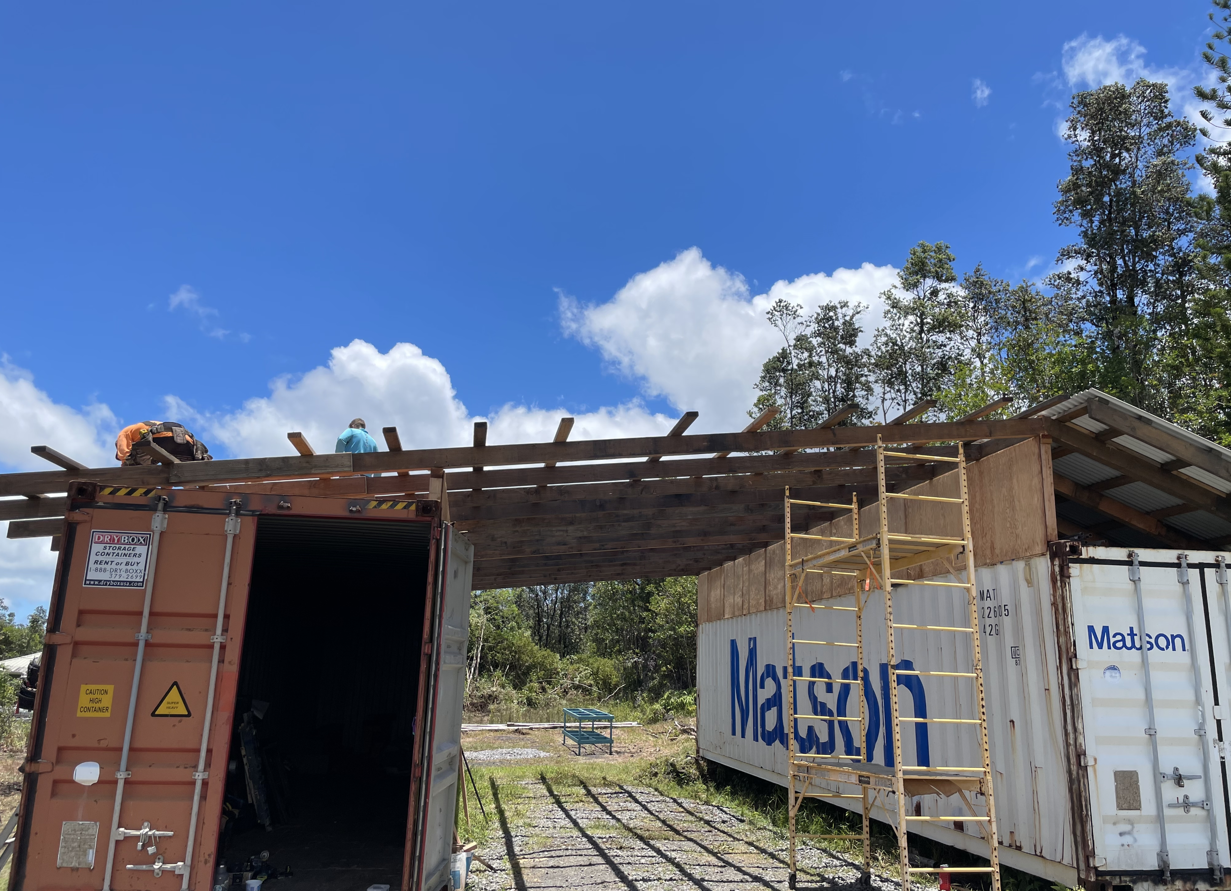 Construction workers building a structure with a clear blue sky and trees in the background, using shipping containers and scaffolding.