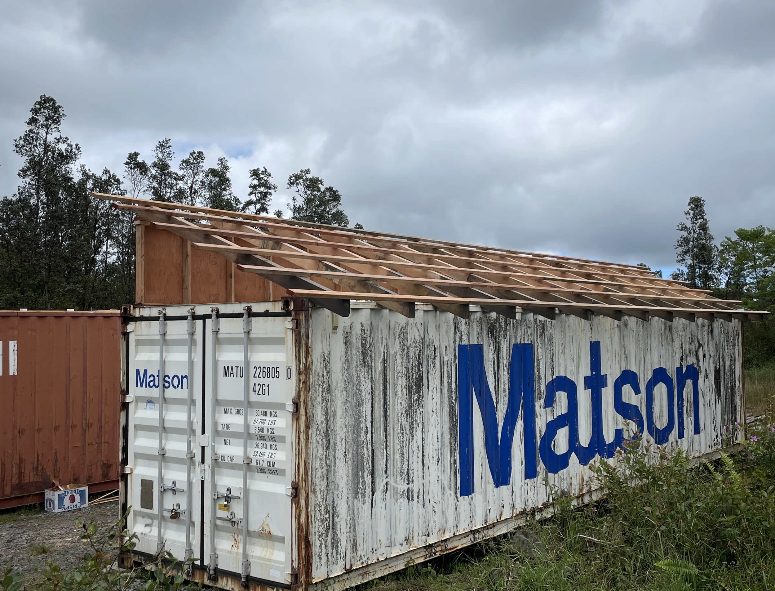 Construction site with a shipping container and a wooden roof framework, surrounded by trees and under a cloudy sky.