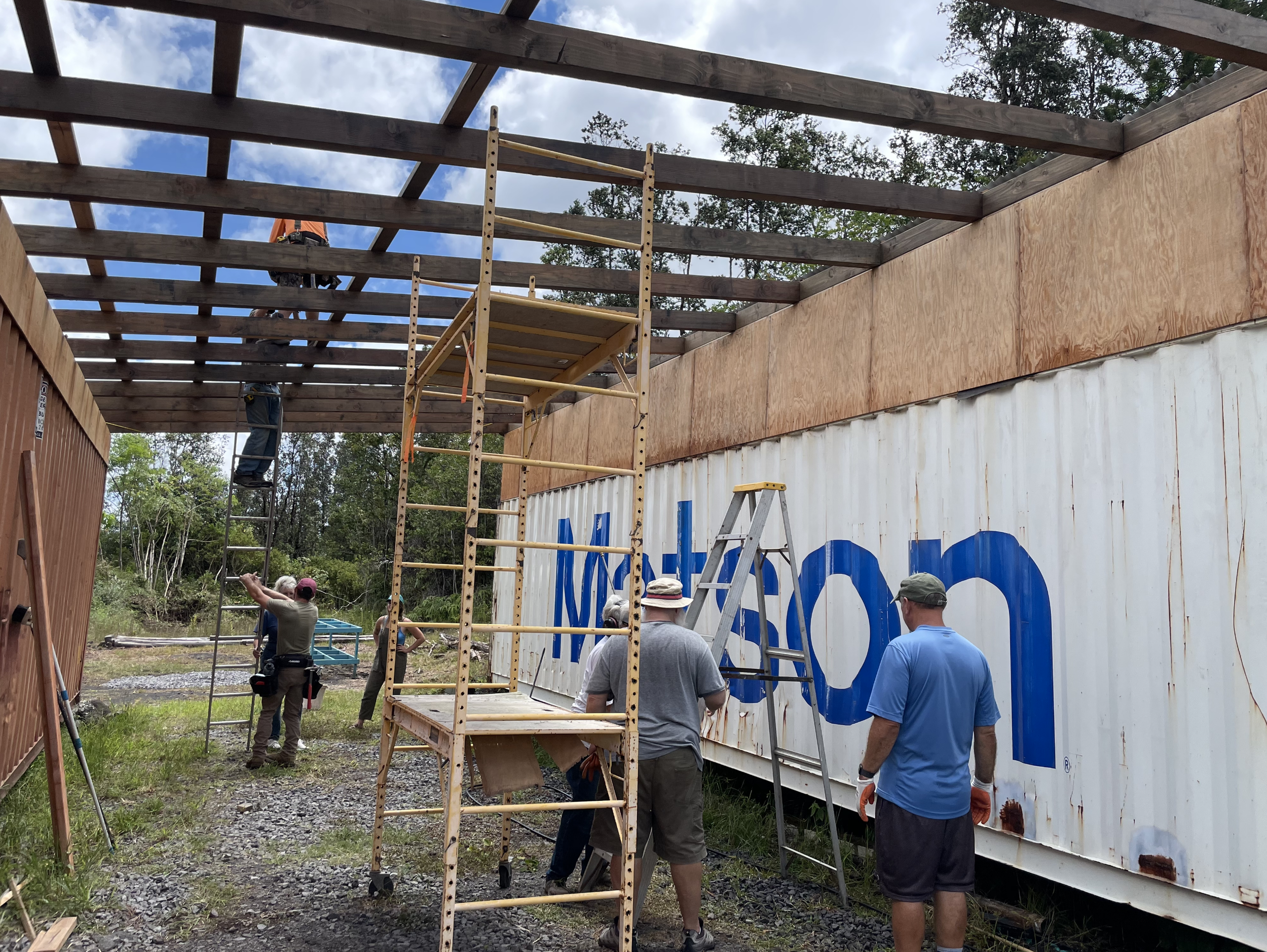 Construction workers are building a roof on a structure with wooden beams and plywood panels, with scaffolding and ladders present at the site.