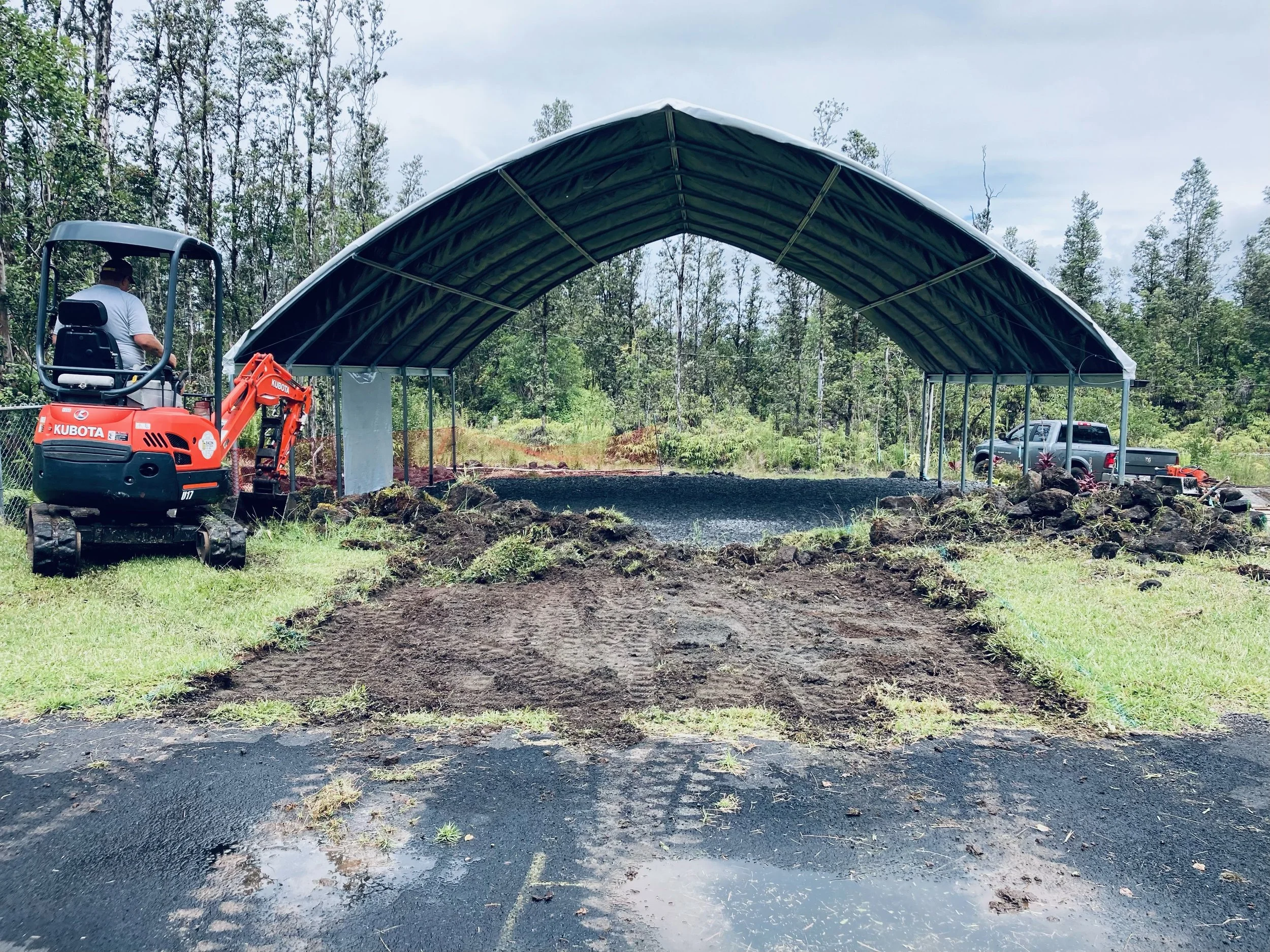 Construction site with a small orange Kubota excavator on the left and a partially built metal structure with a curved roof in the center, surrounded by trees and greenery.