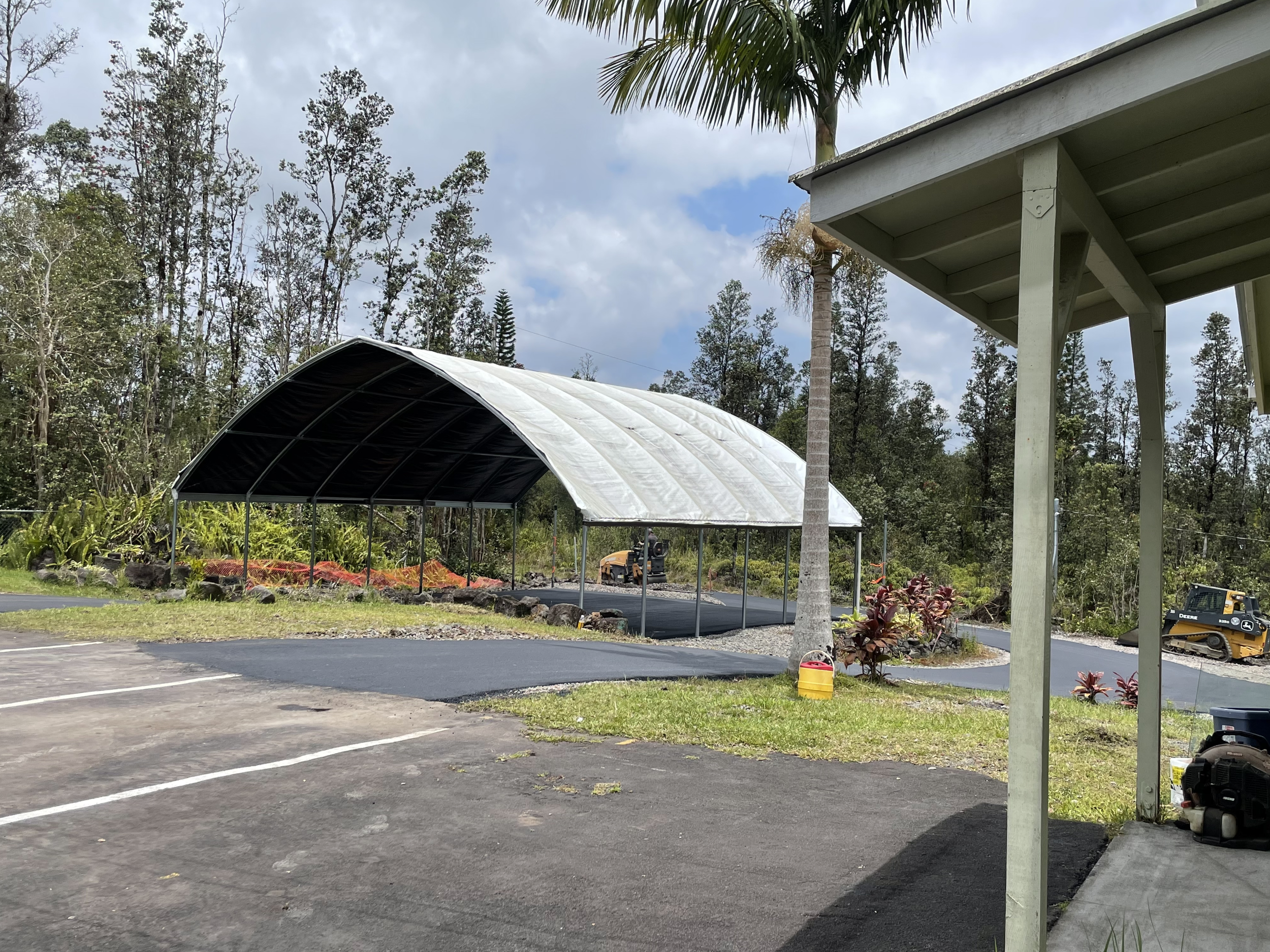 An outdoor parking lot area with a large open-sided shelter and a small building, surrounded by trees and plants.