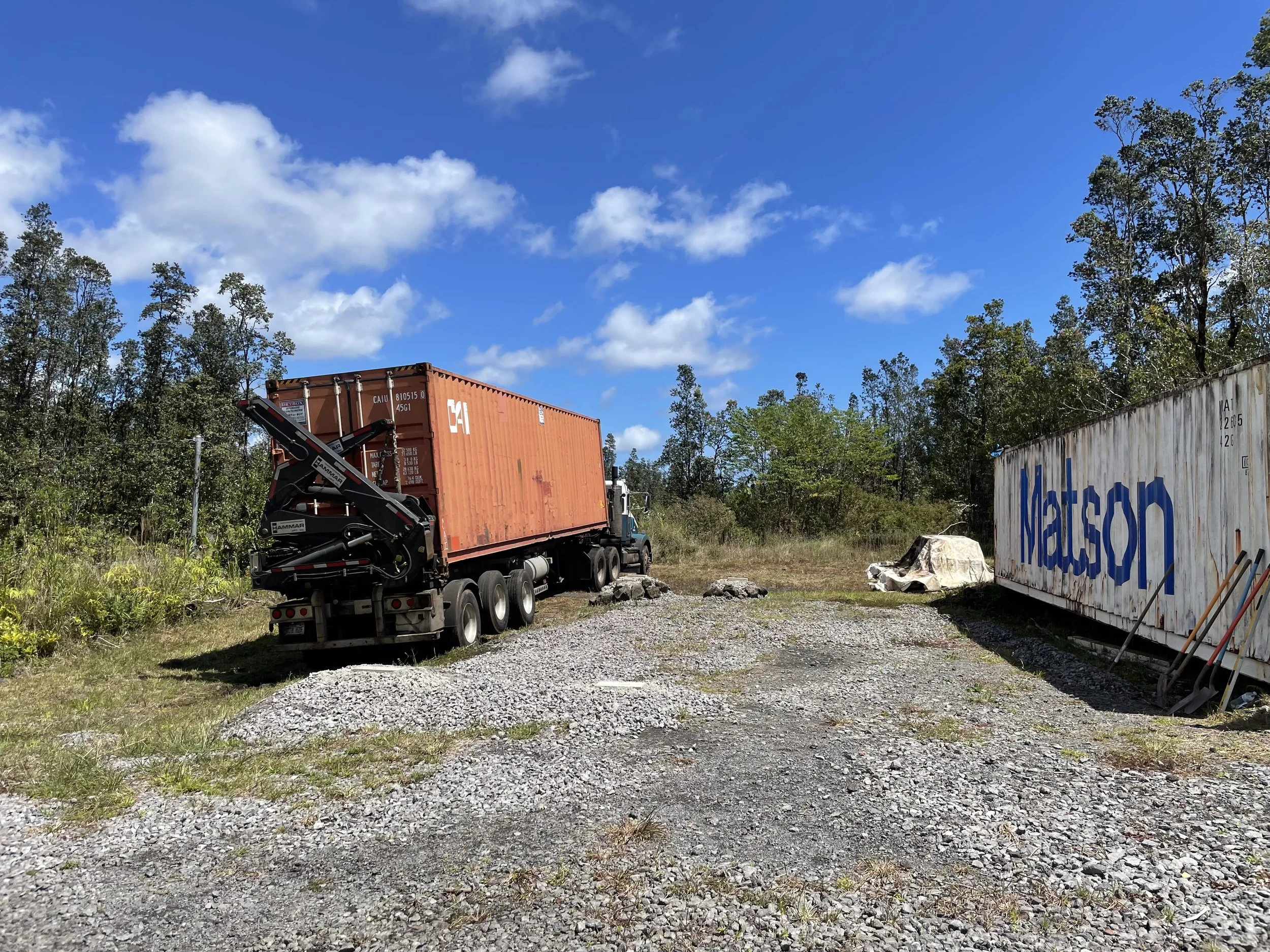 A semi-truck carrying a large orange shipping container parked on a gravel lot surrounded by green trees and blue sky with scattered clouds.