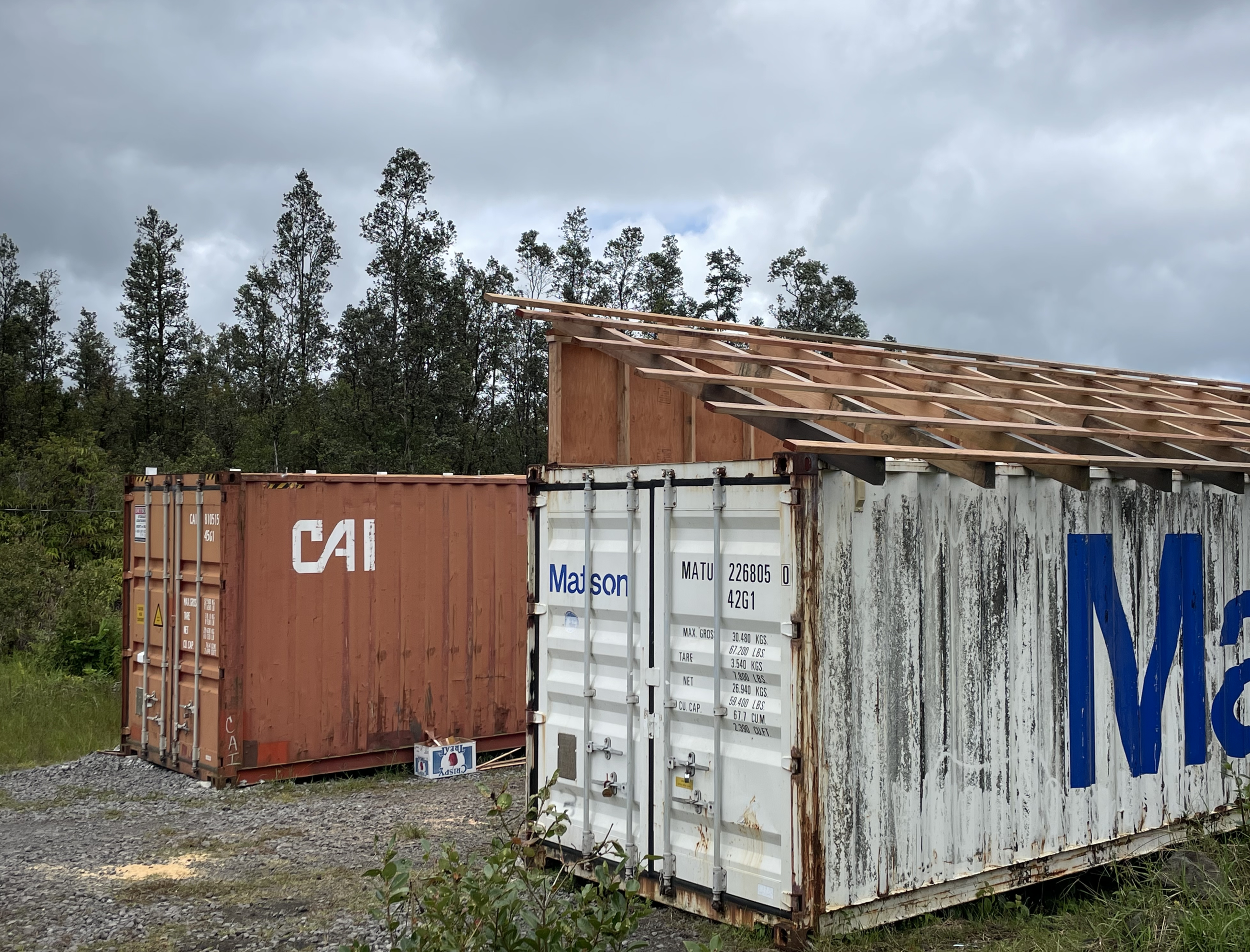 Several shipping containers stacked outdoors near a wooded area, with a partly overcast sky overhead.