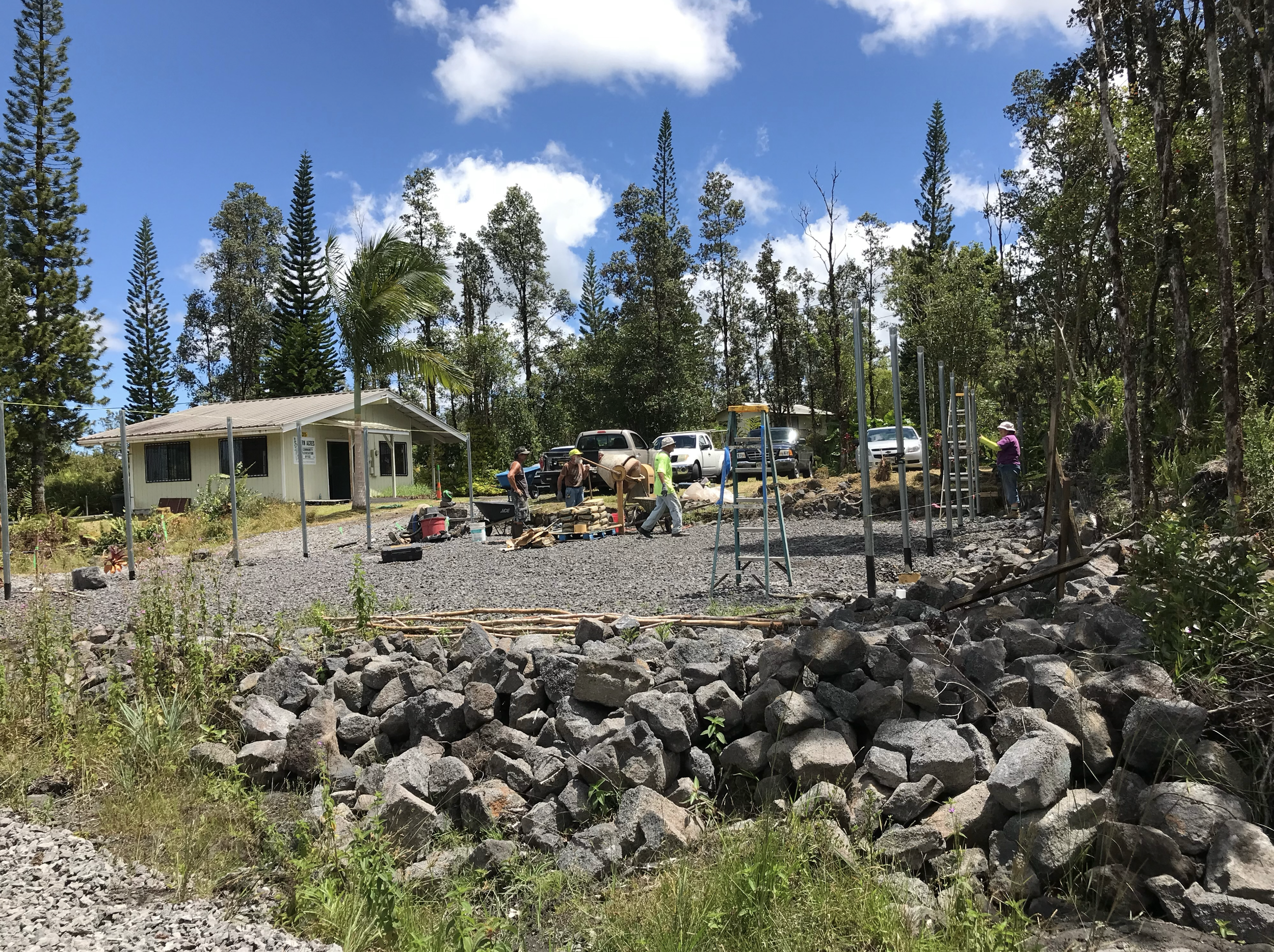 Construction workers building a fence in a rural area with trees, a small building, and cars in the background.