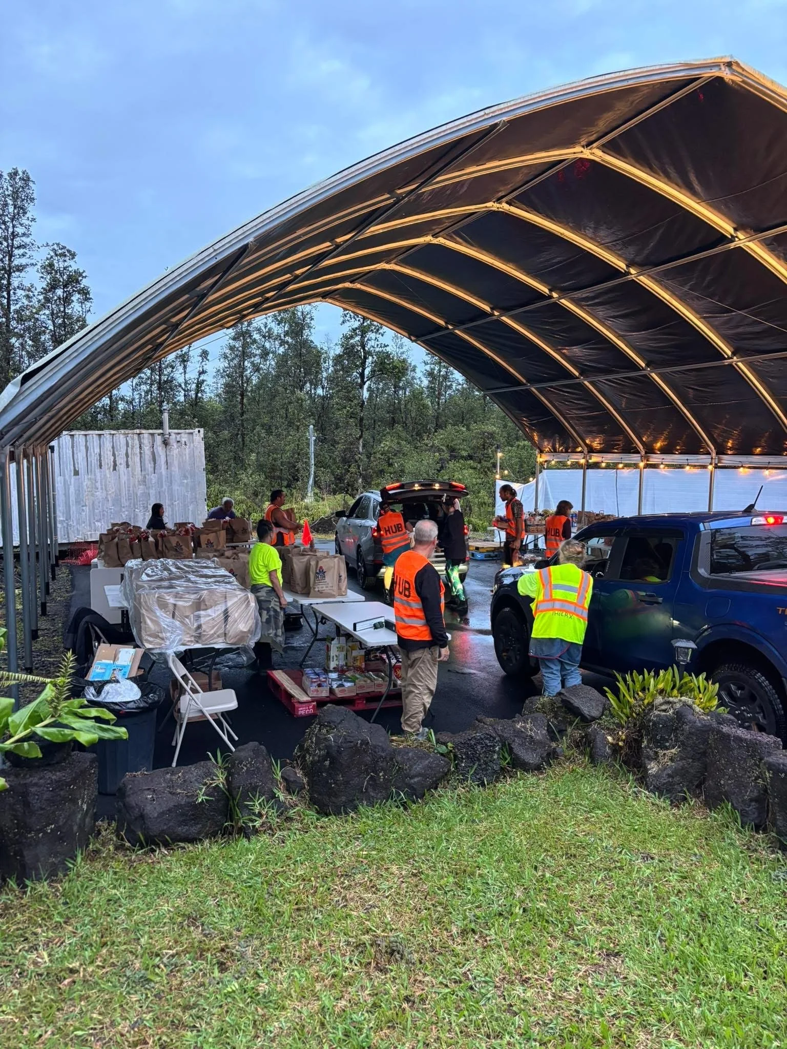 Group of people under a large canopy tent, preparing food and distributing supplies, with parked cars and trees in the background.
